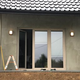 Elektryk 24H - Montaż zewnętrznego oświetlenia na elewacji domu z otwartym oknem balkonowym. Na pierwszym planie drabina i poziomica. Widoczne narzędzia.