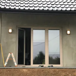 Montaż zewnętrznego oświetlenia na elewacji domu z otwartym oknem balkonowym. Na pierwszym planie drabina i poziomica. Widoczne narzędzia.