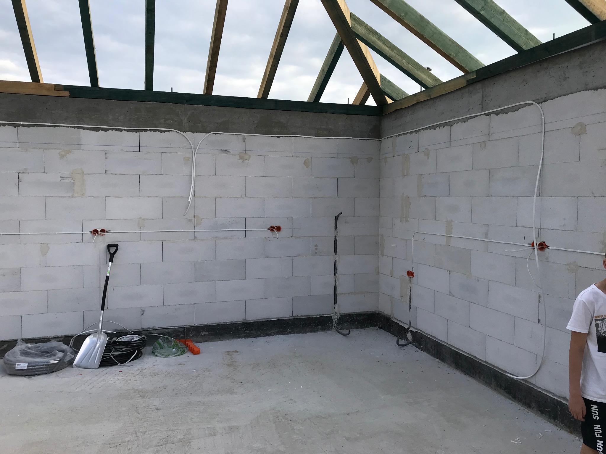 Wnętrze w trakcie budowy z widoczną instalacją elektryczną: kable poprowadzone po ścianach z betonu komórkowego, puszki elektryczne. Widoczne narzędzia i częściowo drewniana konstrukcja dachu.