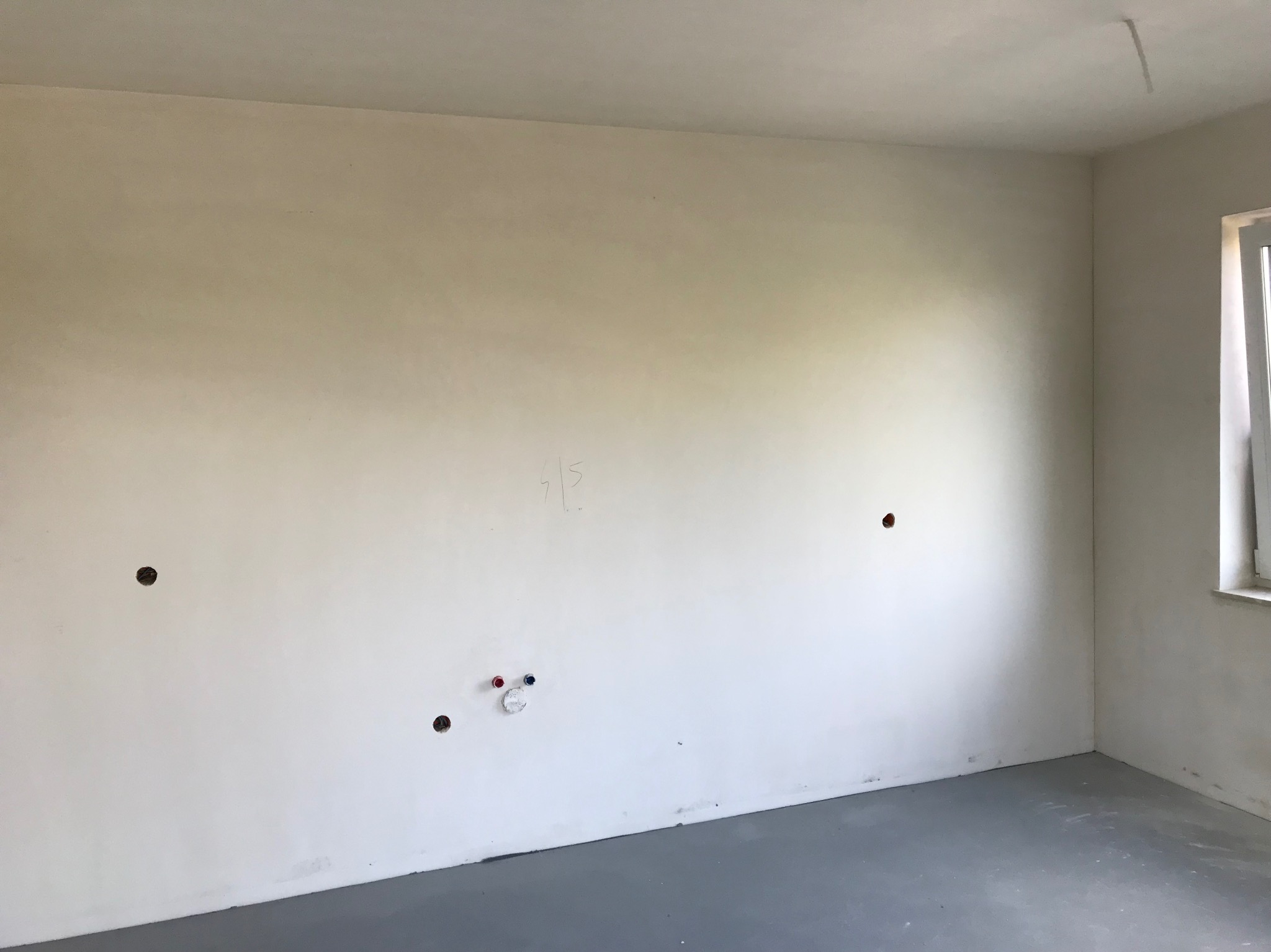 Wnętrze w stanie deweloperskim: gładkie ściany z widocznymi otworami na instalacje elektryczne i wodno-kanalizacyjne, szara wylewka, okno z widocznym fragmentem ramy.