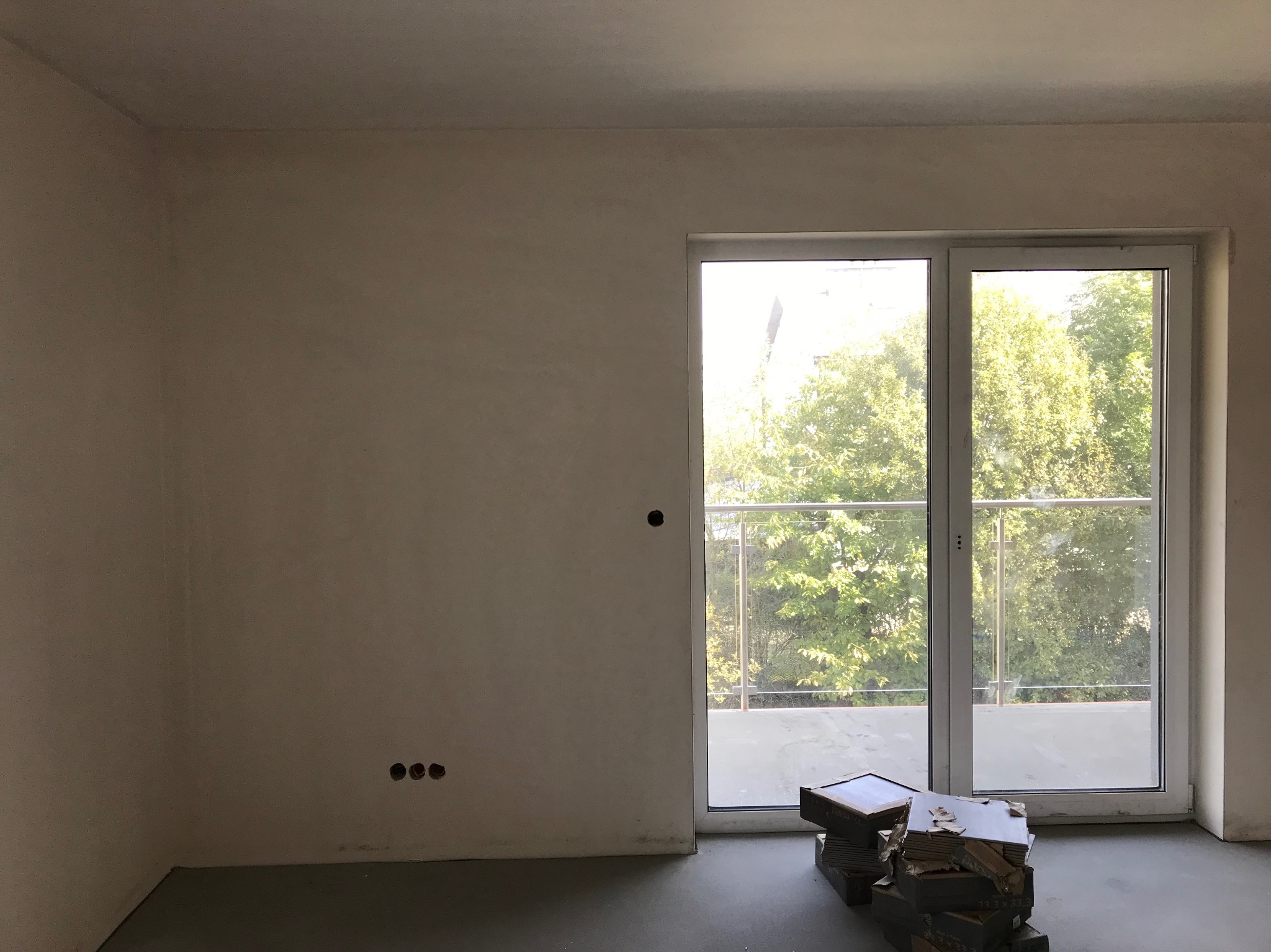 Surowy pokój z balkonem i widocznymi puszkami elektrycznymi w ścianie, przygotowany do prac wykończeniowych. Na podłodze leżą płytki.