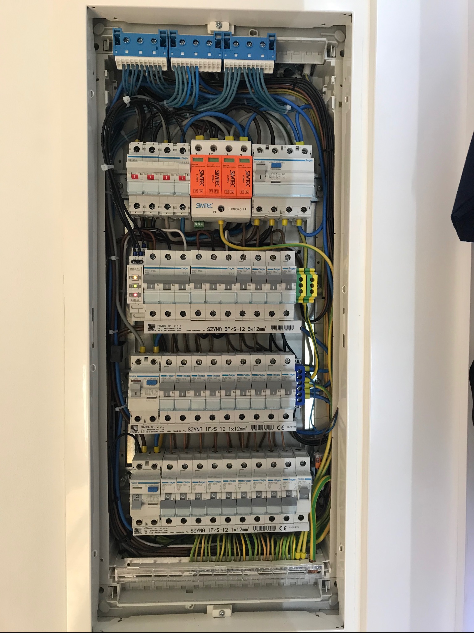 Rozdzielnica elektryczna z bezpiecznikami, przewodami i listwami zaciskowymi. Widoczne elementy marki Hager i Simtec. Całość w białej obudowie.