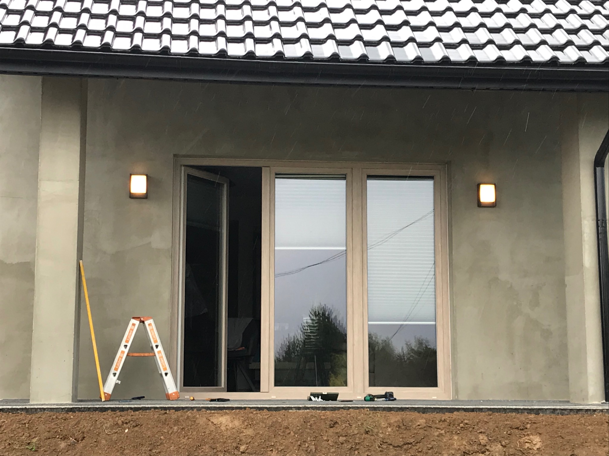 Montaż zewnętrznego oświetlenia na elewacji domu z otwartym oknem balkonowym. Na pierwszym planie drabina i poziomica. Widoczne narzędzia.