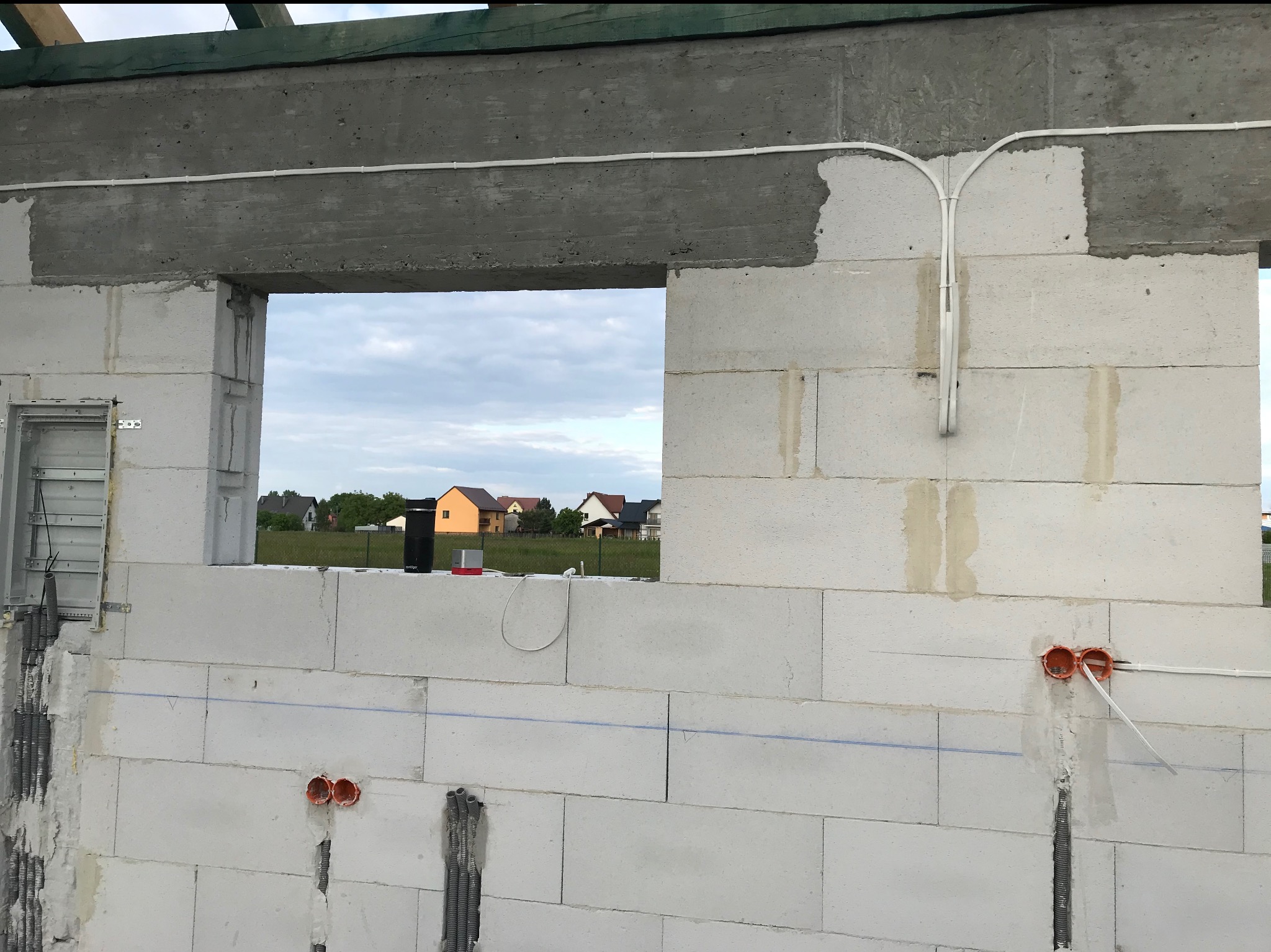 Ściana z betonu komórkowego w trakcie budowy domu, widoczne kable i puszki instalacyjne. Przez okno widać zabudowania i niebo.