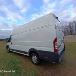 Biały, dostawczy bus na trawiastym terenie, widok z tyłu, z widoczną tablicą rejestracyjną. Pochmurne niebo w tle.