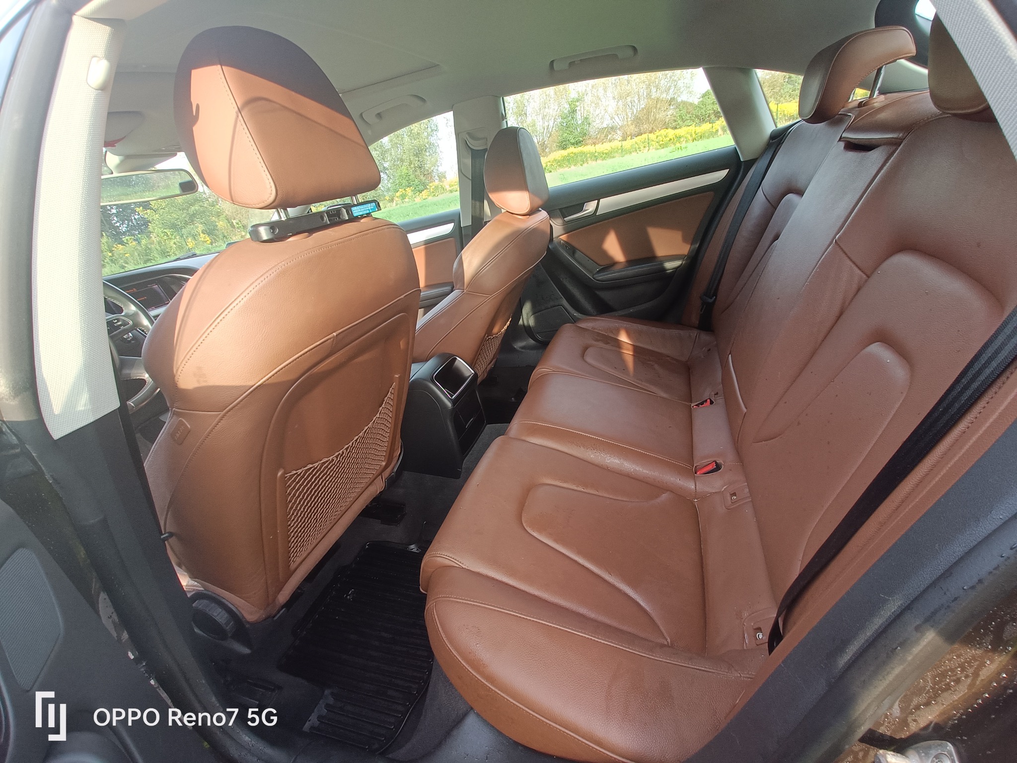 Wnętrze samochodu z brązową skórzaną tapicerką, widok na tylne siedzenia. Widoczne zagłówki, pasy bezpieczeństwa i siatka na oparciu przedniego fotela. Widok z perspektywy tylnych drzwi.