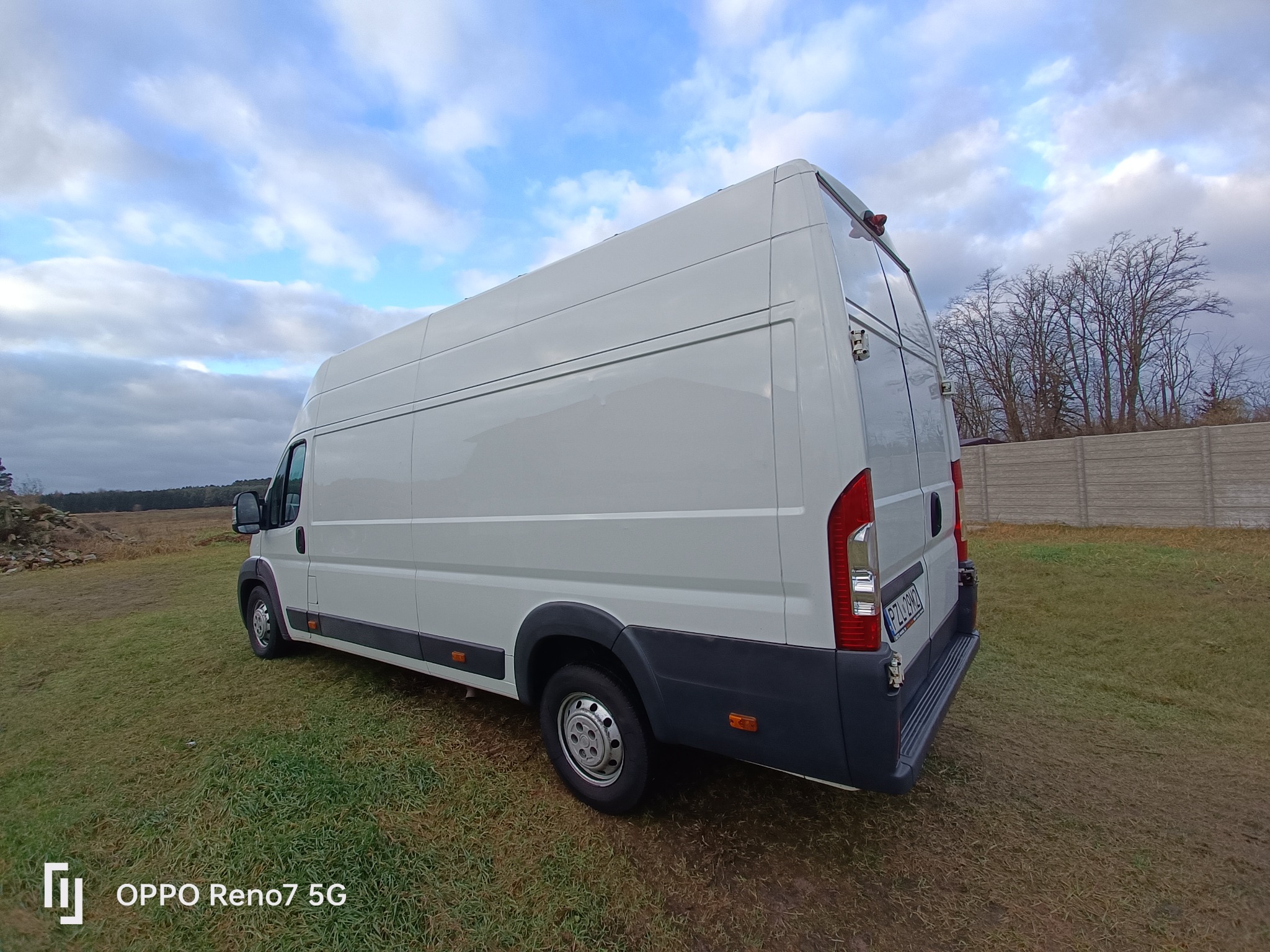 Biały, dostawczy bus na trawiastym terenie, widok z tyłu, z widoczną tablicą rejestracyjną. Pochmurne niebo w tle.