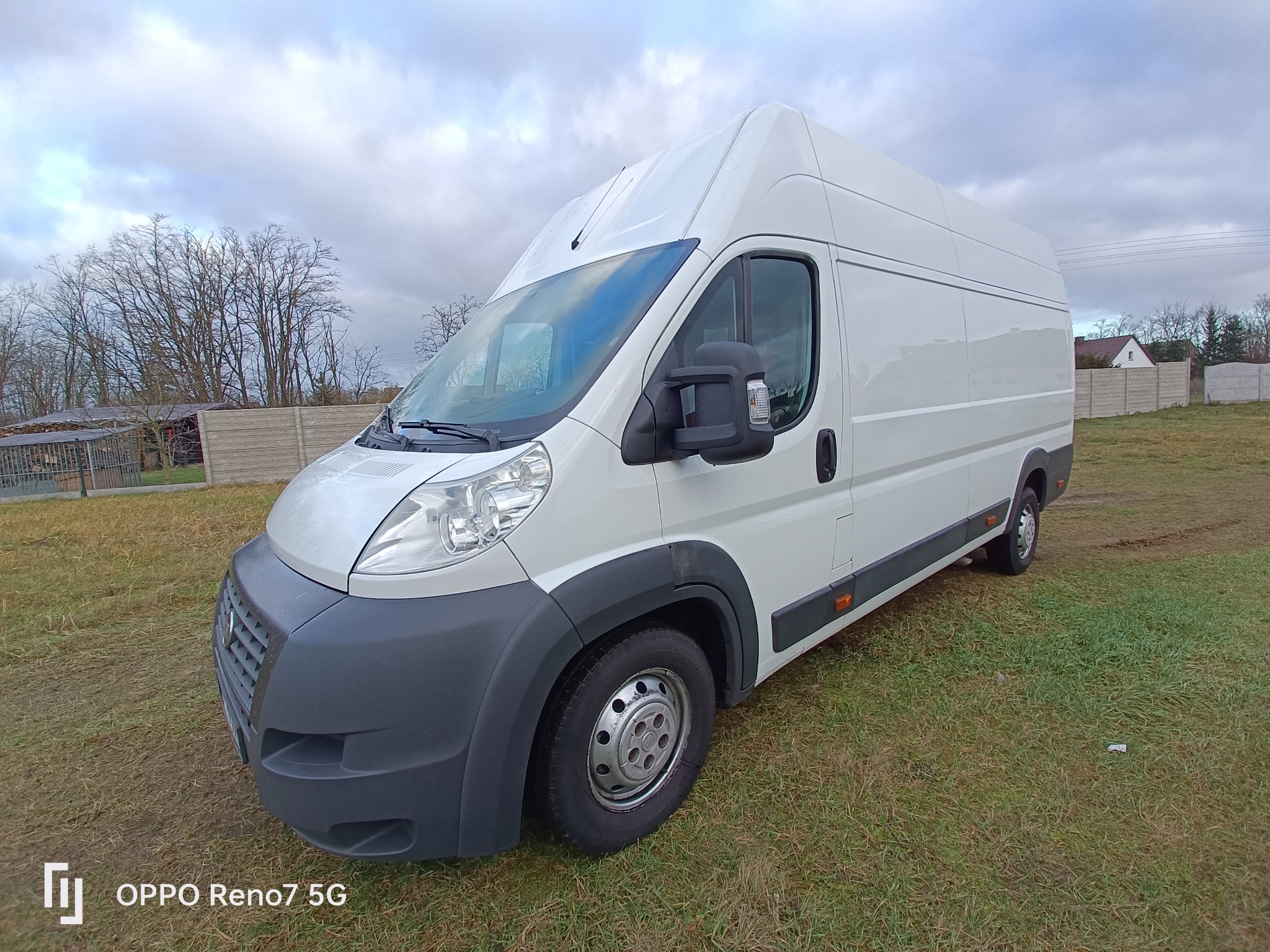 Biały, dostawczy bus marki Fiat Ducato, sfotografowany pod kątem na trawiastym terenie z widocznym ogrodzeniem betonowym w tle, pochmurne niebo.