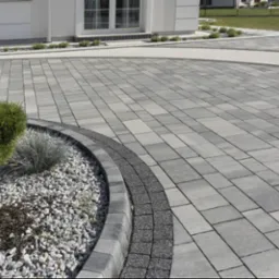 Szara kostka brukowa ułożona na podjeździe z dekoracyjnym obramowaniem i rabatą z kamykami oraz roślinami, przylegająca do budynku z białymi elementami architektonicznymi.