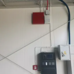 Panel sterowania alarmem pożarowym Bosch na białej ścianie z widocznymi przewodami i elementami instalacji w przemysłowym otoczeniu.