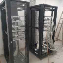 Firma Elektryczna D-Volt Daniel Kruczek - Dwie szafy serwerowe w trakcie instalacji okablowania, widoczne kable i drabina w tle. Prace instalacyjne w pomieszczeniu z białymi ścianami i podwieszanym sufitem.