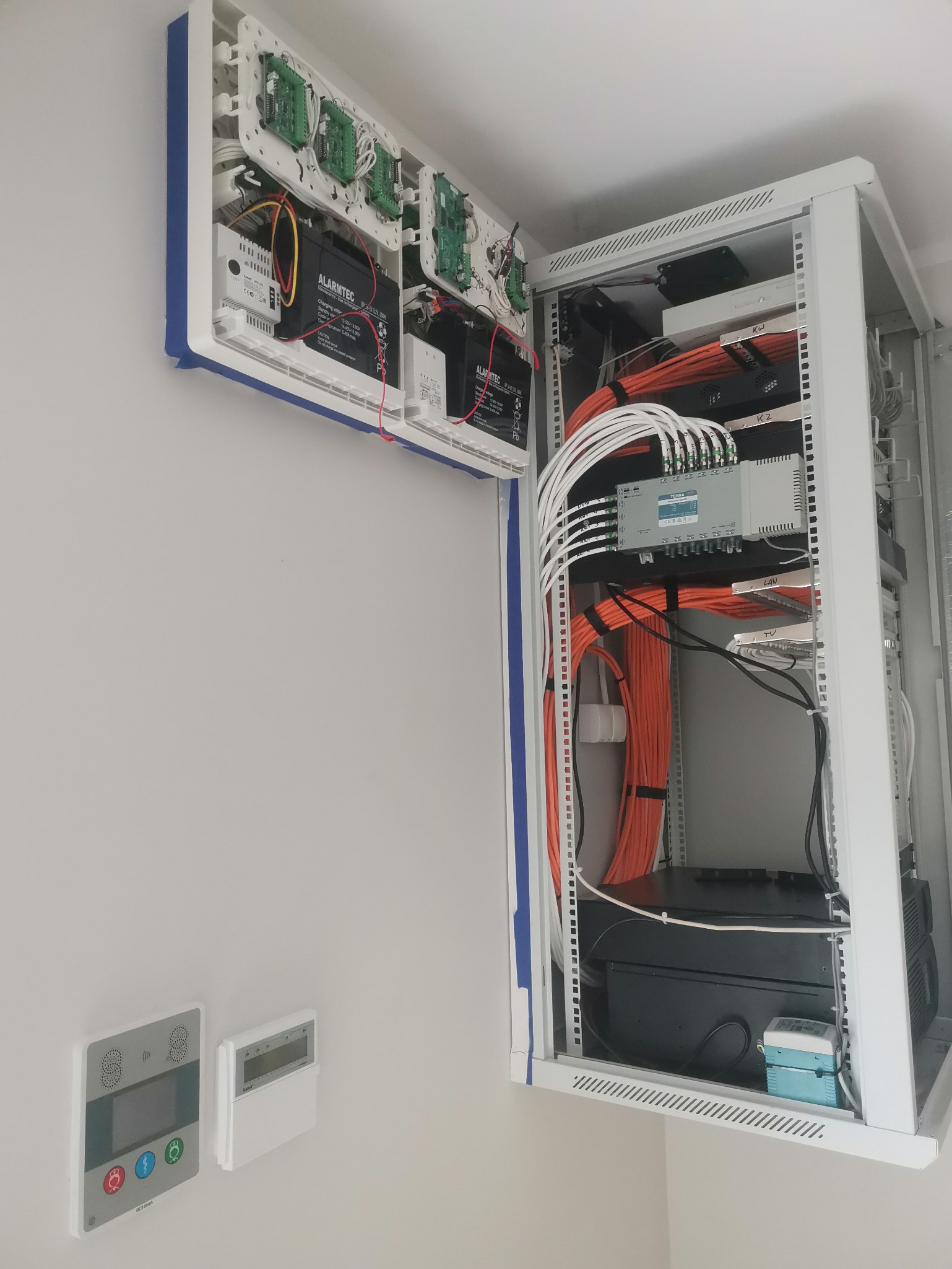 Rozdzielnia alarmowa z akumulatorami i okablowaniem w szafie rack, panele sterujące alarmem na ścianie. Widok na zainstalowany system zabezpieczeń.