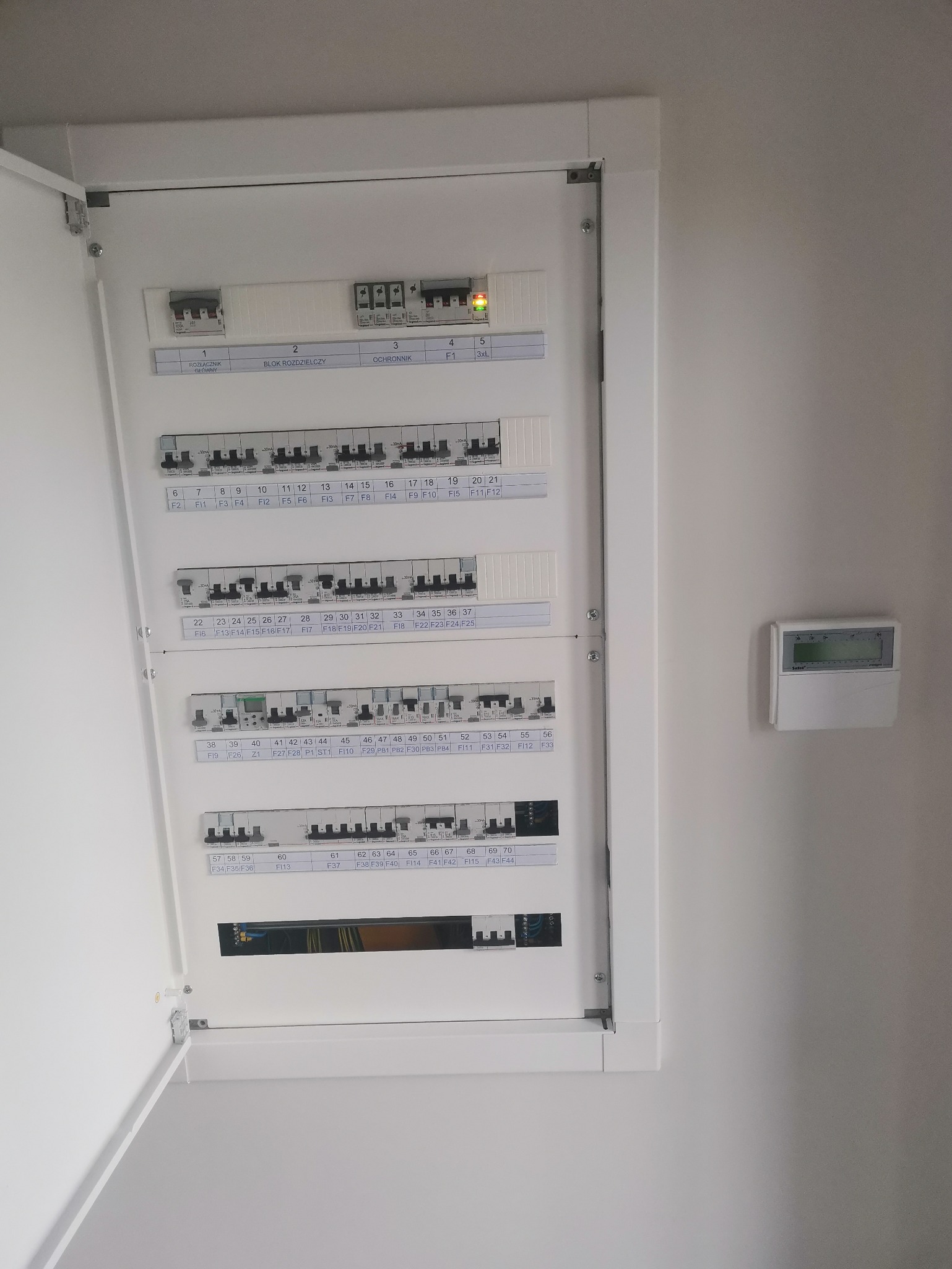 Otwarta skrzynka elektryczna z widocznymi bezpiecznikami i oznaczeniami wewnątrz białej obudowy, zamontowana na jasnej ścianie wewnątrz pomieszczenia.