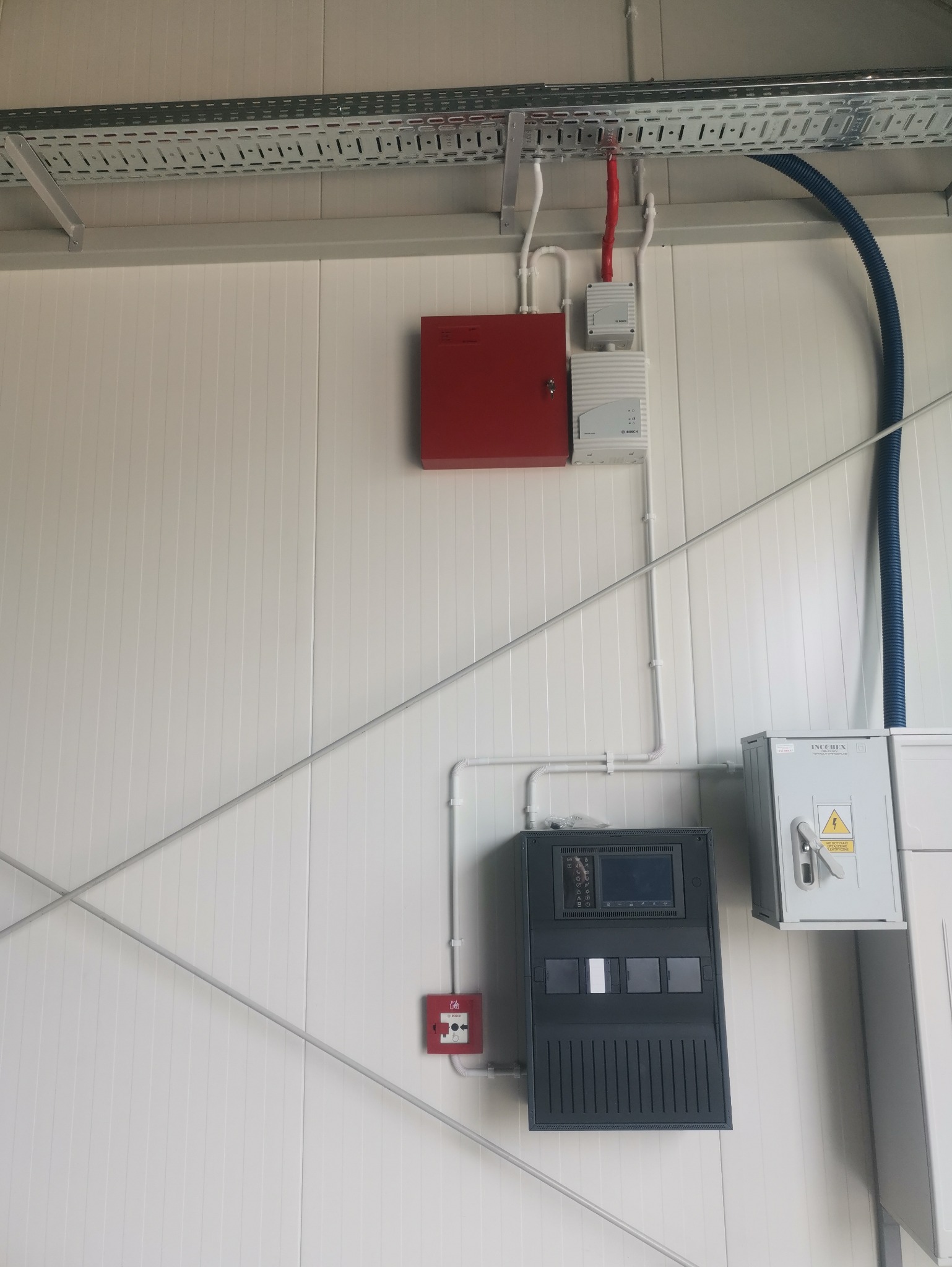 Panel sterowania alarmem pożarowym Bosch na białej ścianie z widocznymi przewodami i elementami instalacji w przemysłowym otoczeniu.