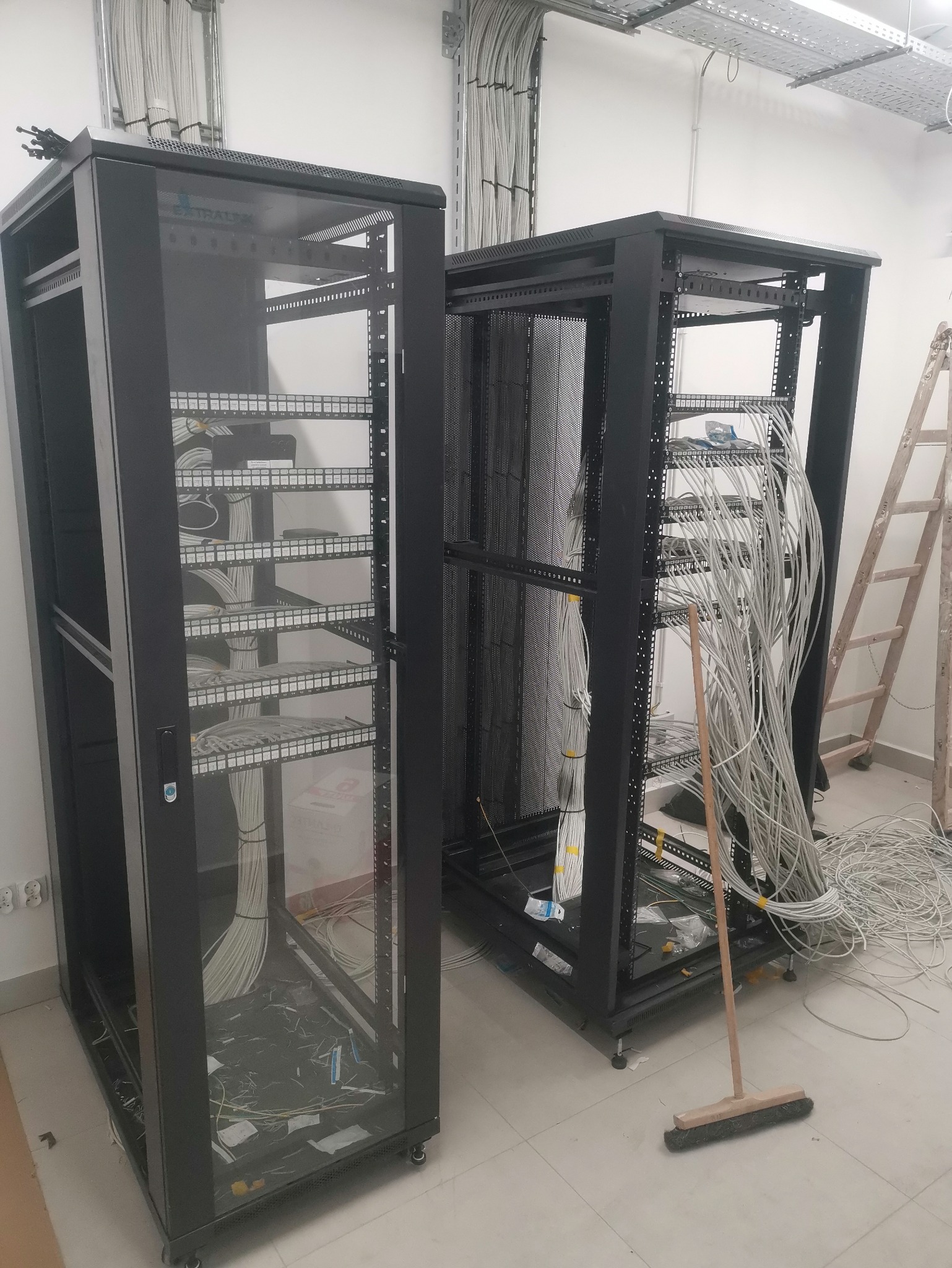 Dwie szafy serwerowe w trakcie instalacji okablowania, widoczne kable i drabina w tle. Prace instalacyjne w pomieszczeniu z białymi ścianami i podwieszanym sufitem.