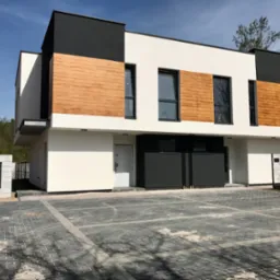 Nowoczesny, dwukondygnacyjny dom z elementami drewna na elewacji, czarnymi akcentami i kostką brukową na podjeździe w słoneczny dzień. Widoczna skrzynka gazowa.