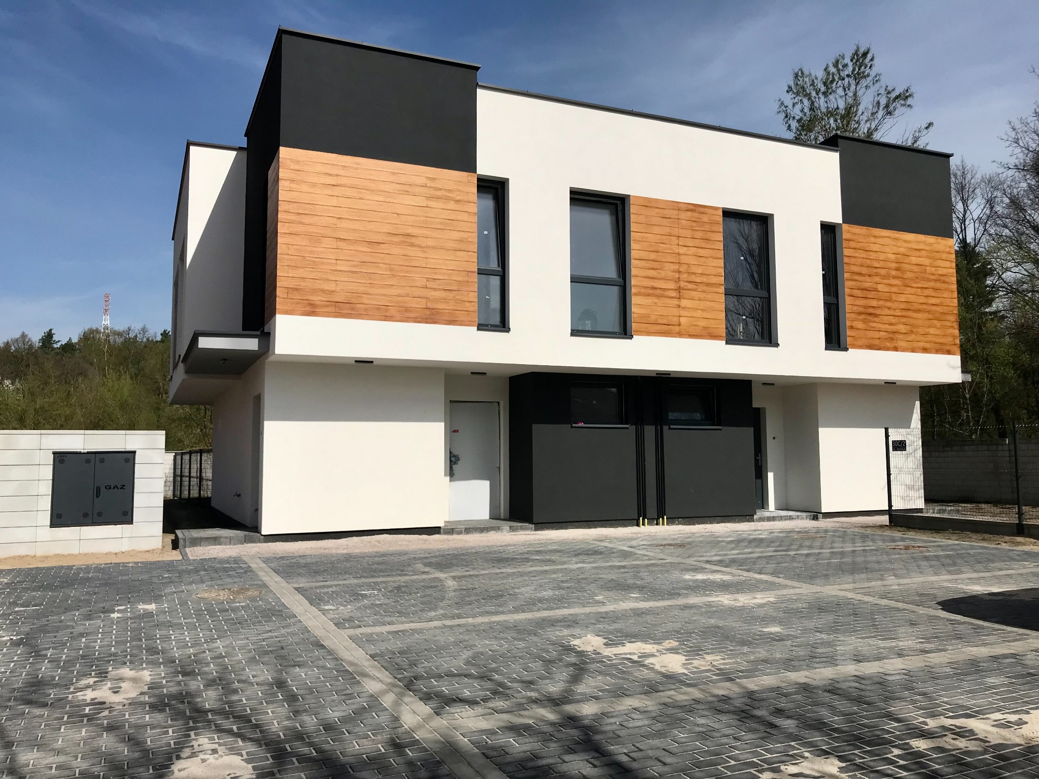Nowoczesny, dwukondygnacyjny dom z elementami drewna na elewacji, czarnymi akcentami i kostką brukową na podjeździe w słoneczny dzień. Widoczna skrzynka gazowa.