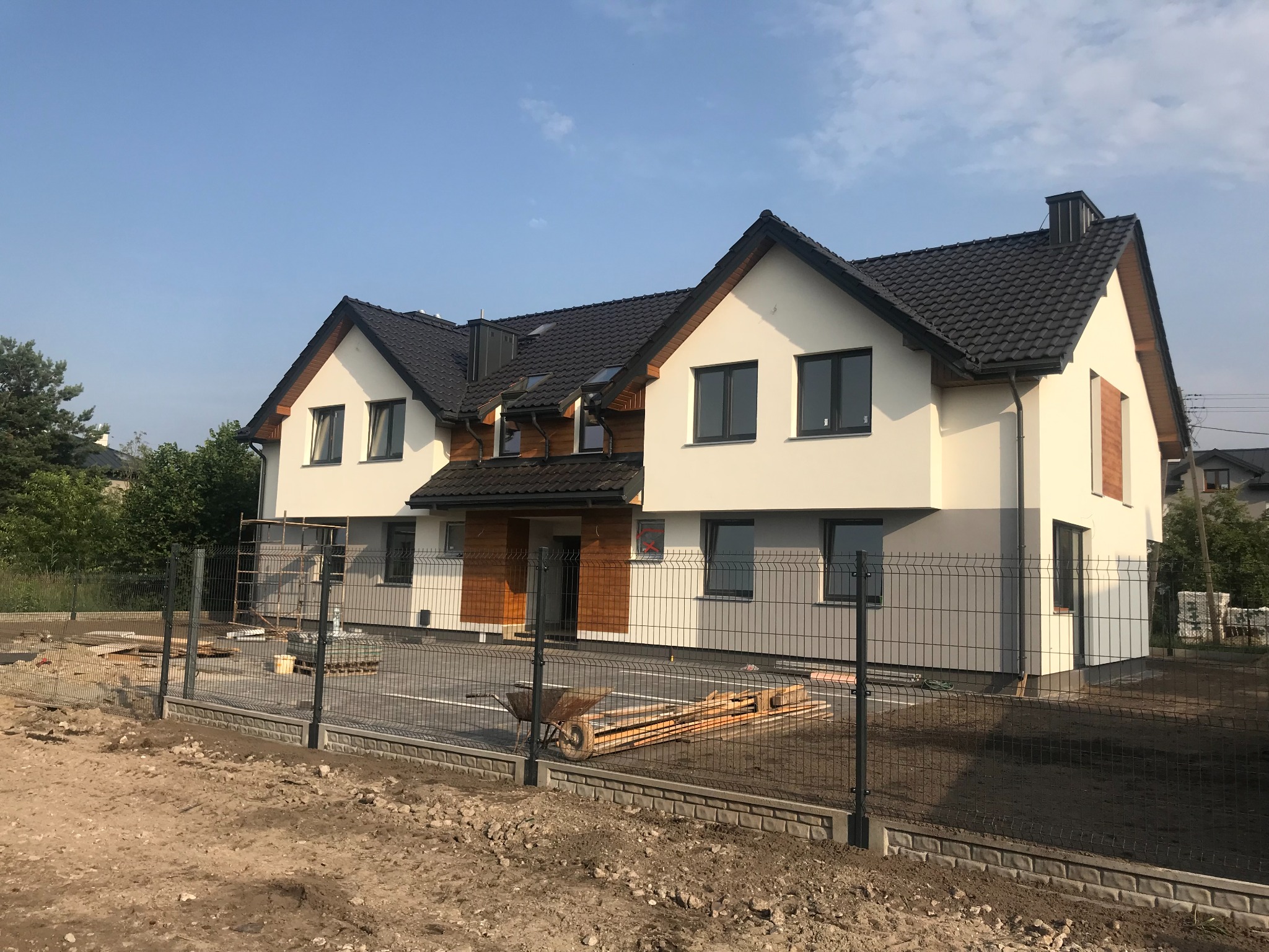 Nowoczesny dom dwukondygnacyjny z ciemną dachówką, jasną elewacją i drewnianymi akcentami. Wokół posesji tymczasowe ogrodzenie panelowe. Widoczne materiały budowlane.