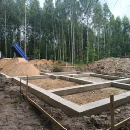 Wylany fundament pod dom na działce z drzewami w tle. Widoczny wywrotka z piaskiem. Świeży beton, szalunki i wykopana ziemia.
