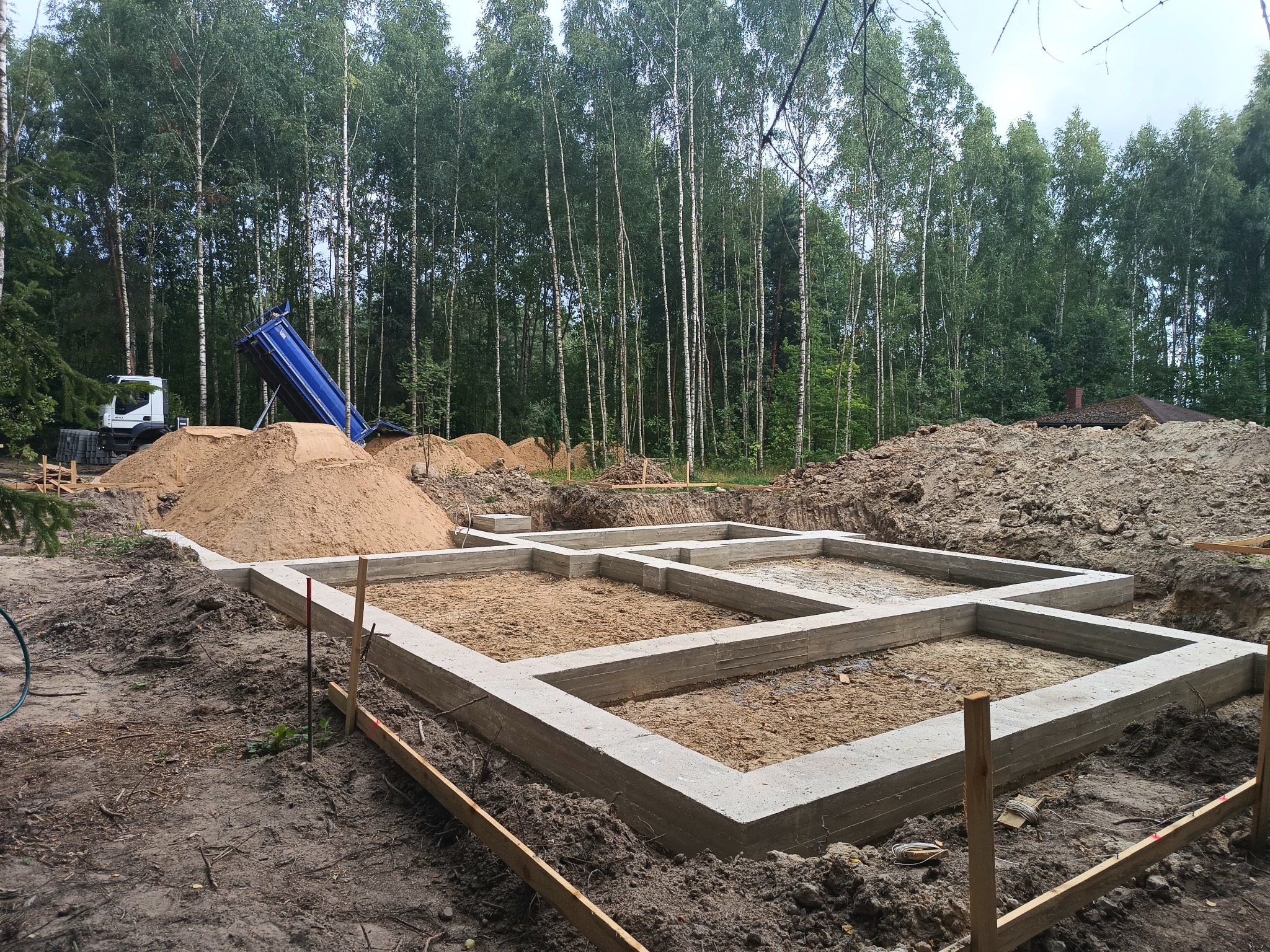 Wylany fundament pod dom na działce z drzewami w tle. Widoczny wywrotka z piaskiem. Świeży beton, szalunki i wykopana ziemia.