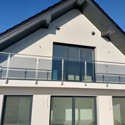 Elewacja domu z balkonem zabezpieczonym szklaną balustradą, widoczne okna z ciemnymi ramami i fragment dachu w słoneczny dzień. Detal architektoniczny.