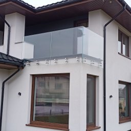 INOX-STYL Mateusz Gabara - Szklana balustrada balkonowa, mocowana punktowo do ściany budynku o białej elewacji, z widocznymi oknami i brązowymi ramami, oraz ciemnym dachem.