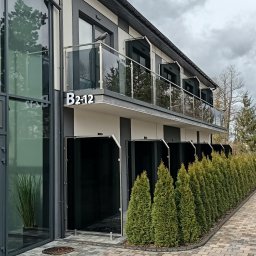 INOX-STYL Mateusz Gabara - Elegancki budynek z balkonami zabezpieczonymi szkłem, widok z zewnątrz, zieleń w tle, nowoczesna architektura, numer B2-12 na elewacji.