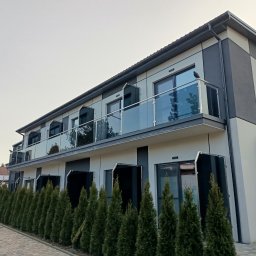 INOX-STYL Mateusz Gabara - Dwupiętrowy budynek z balkonami zabezpieczonymi szkłem, widok z dołu. Rząd tuj przed budynkiem, szaro-biała elewacja. Ptak siedzi na balustradzie.