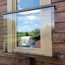 Szklana zabudowa balkonu w drewnianym domu, widok z zewnątrz, z refleksami otoczenia. Detal mocowania szkła do drewnianej konstrukcji.