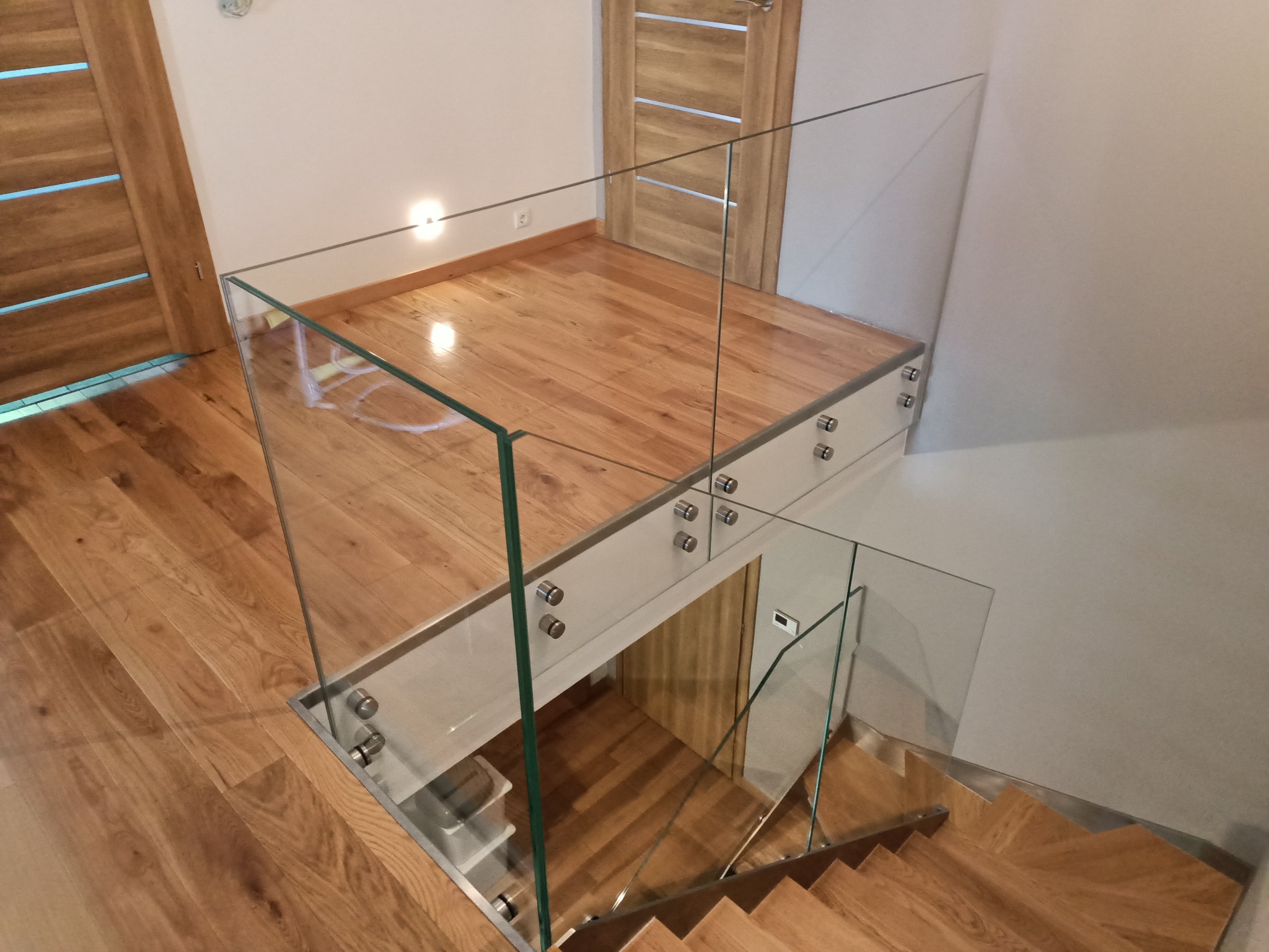 Szklana balustrada z metalowymi mocowaniami, zabezpieczająca schody w jasnym wnętrzu z drewnianą podłogą i drzwiami. Widok z góry na konstrukcję.