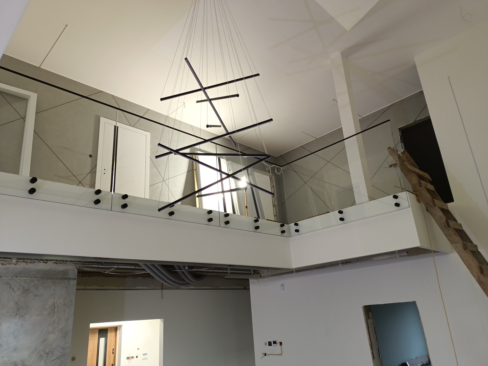 Nowoczesna balustrada szklana z czarnymi mocowaniami na antresoli w surowym wnętrzu, widok z dołu na minimalistyczną lampę wiszącą i fragment drewnianej drabiny.