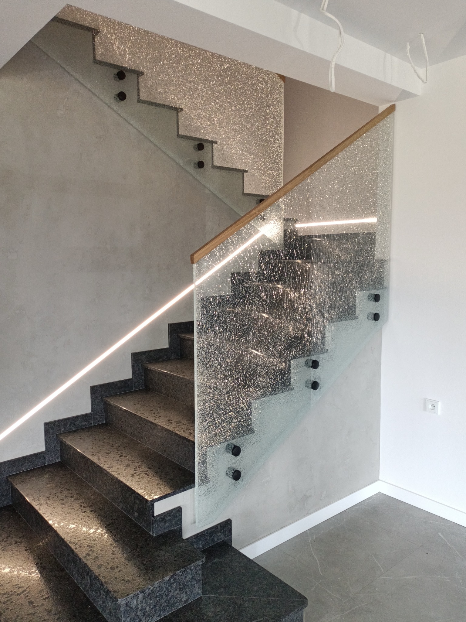 Schody z ciemnego kamienia z balustradą ze szkła ornamentowego i drewnianą poręczą, oświetlone listwą LED. Widok z dołu, podkreślający nowoczesny design i detale wykończenia.