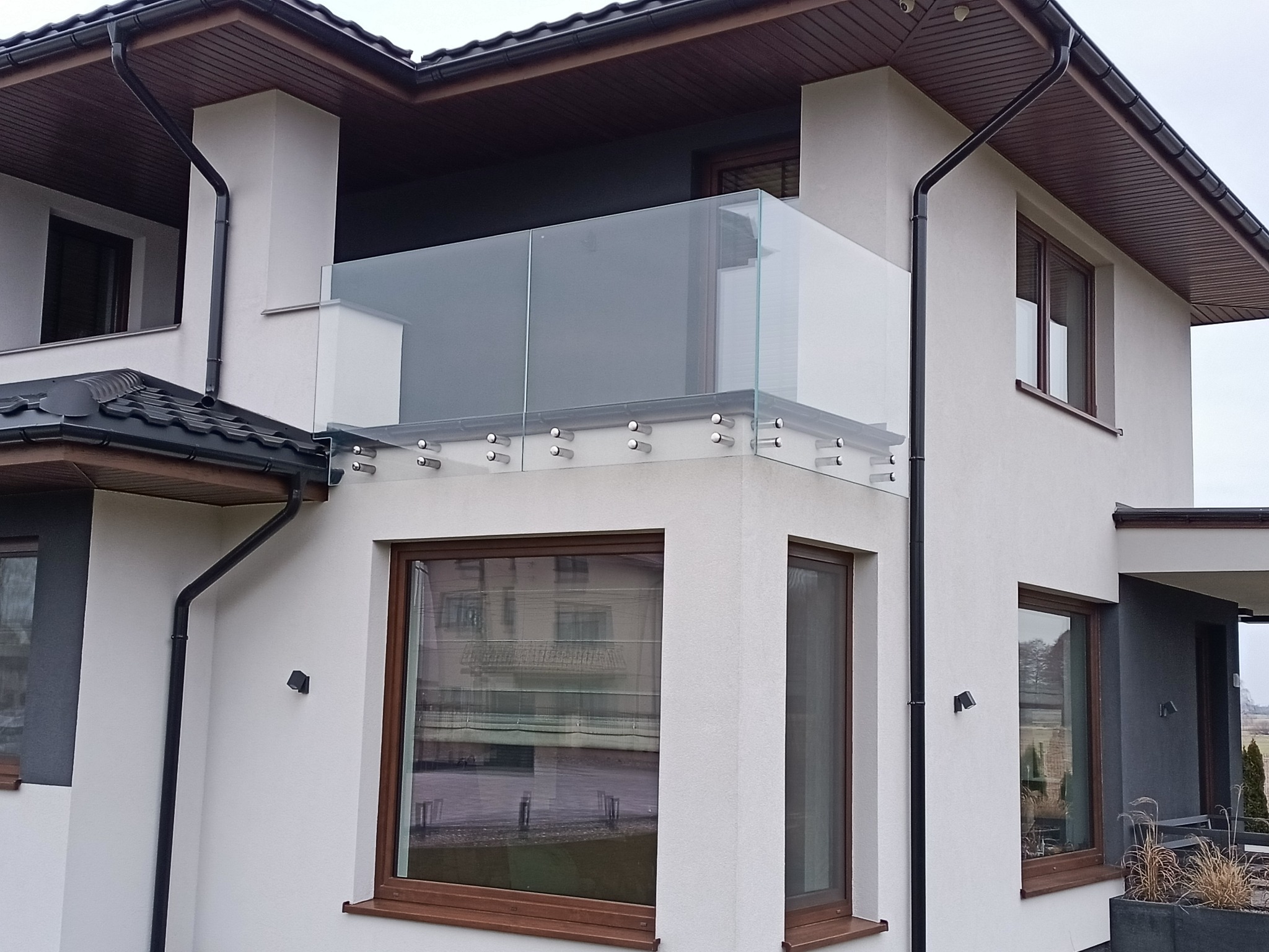Szklana balustrada balkonowa, mocowana punktowo do ściany budynku o białej elewacji, z widocznymi oknami i brązowymi ramami, oraz ciemnym dachem.