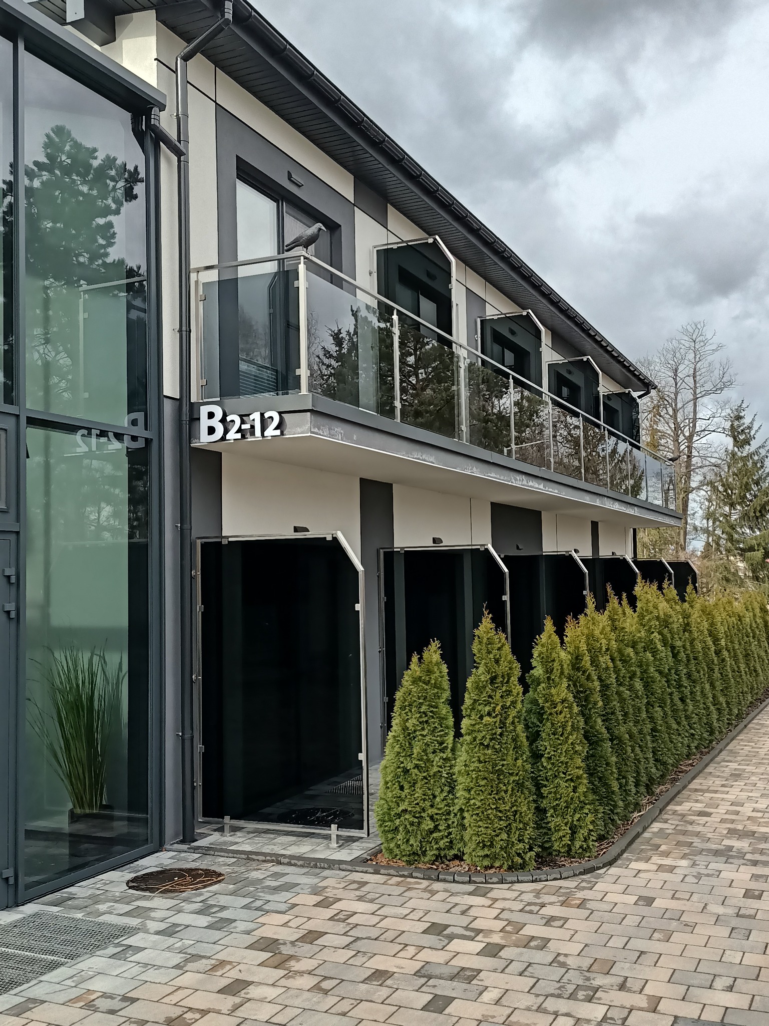 Elegancki budynek z balkonami zabezpieczonymi szkłem, widok z zewnątrz, zieleń w tle, nowoczesna architektura, numer B2-12 na elewacji.