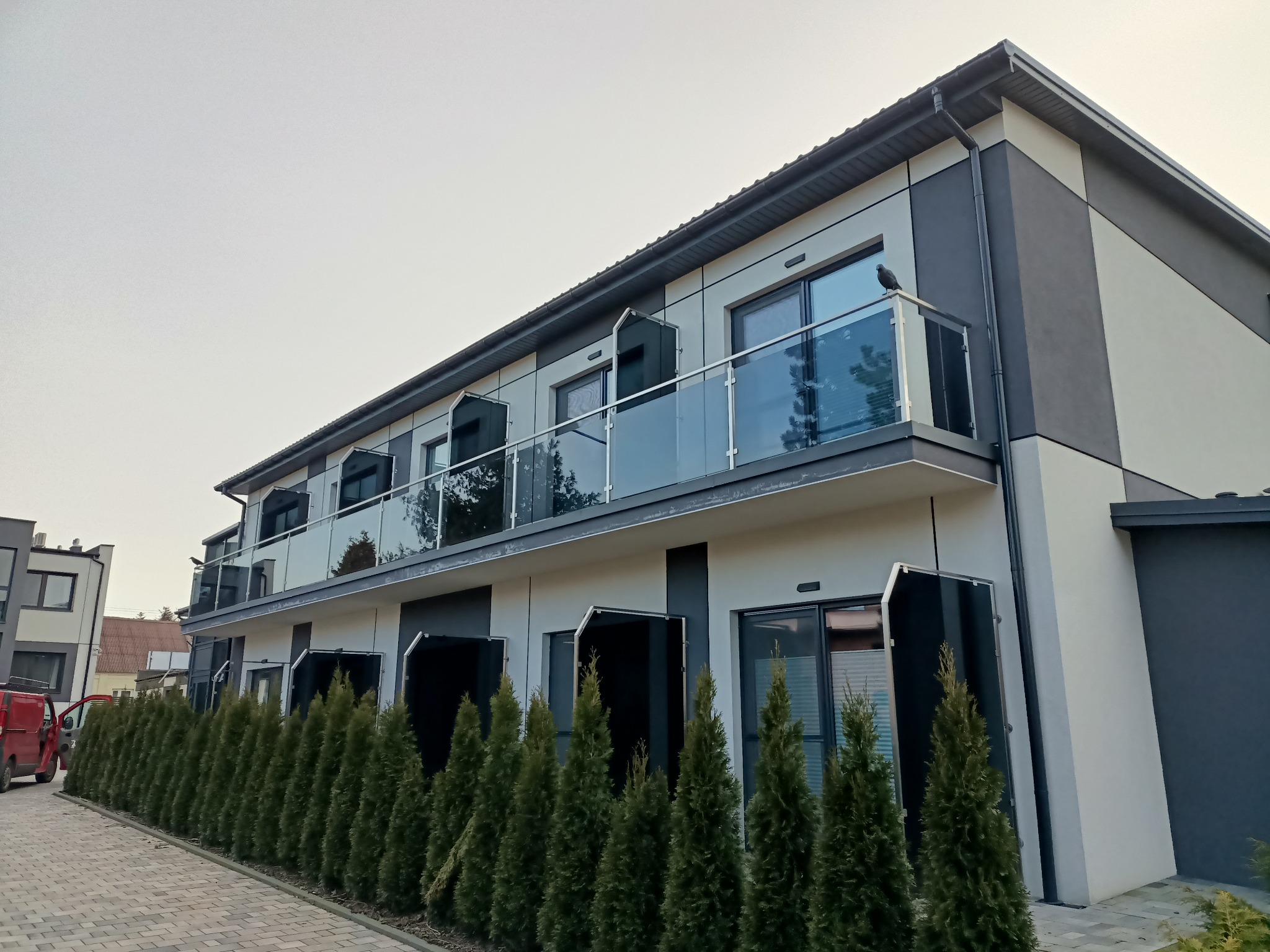 Dwupiętrowy budynek z balkonami zabezpieczonymi szkłem, widok z dołu. Rząd tuj przed budynkiem, szaro-biała elewacja. Ptak siedzi na balustradzie.