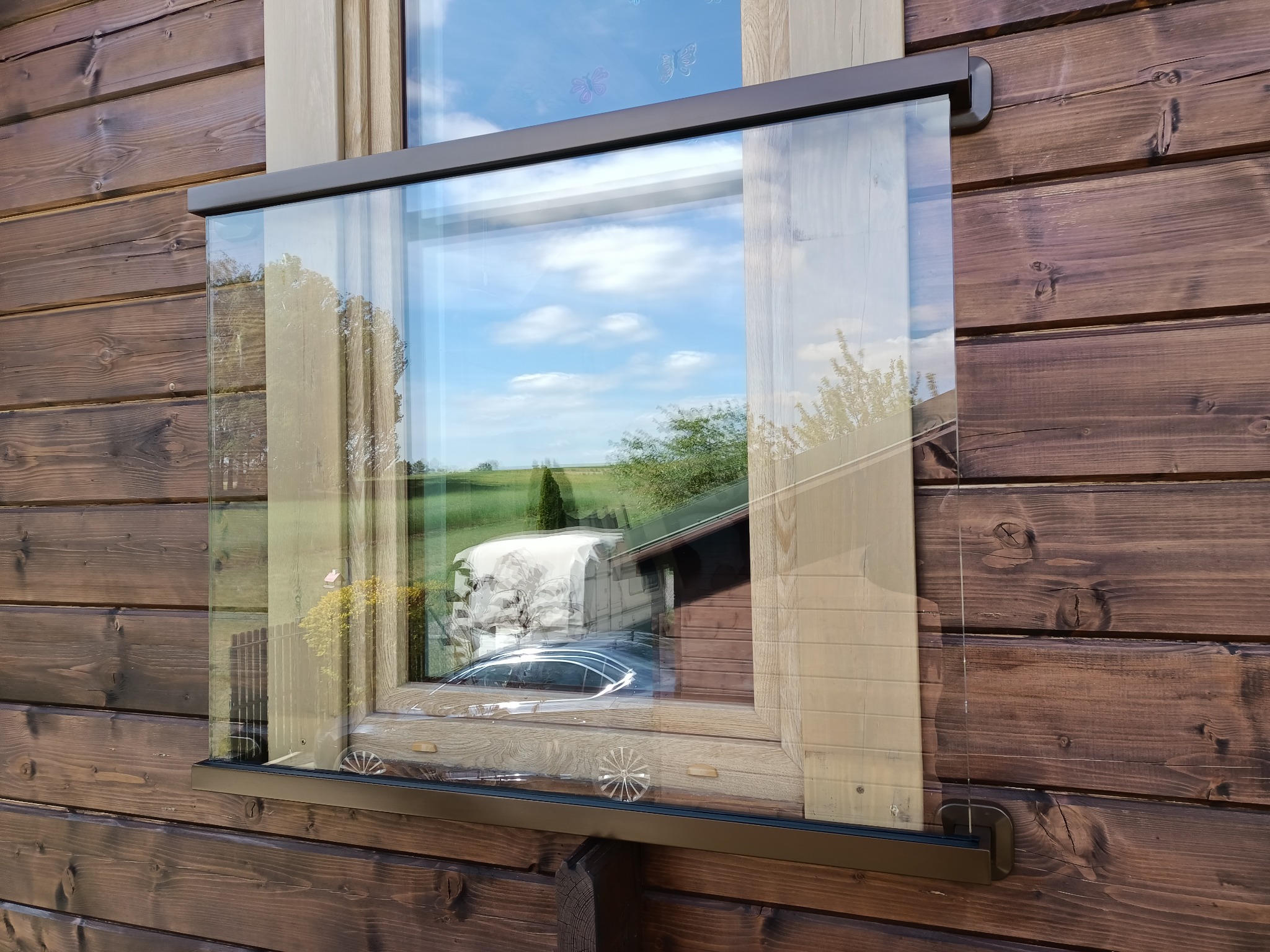 Szklana zabudowa balkonu w drewnianym domu, widok z zewnątrz, z refleksami otoczenia. Detal mocowania szkła do drewnianej konstrukcji.