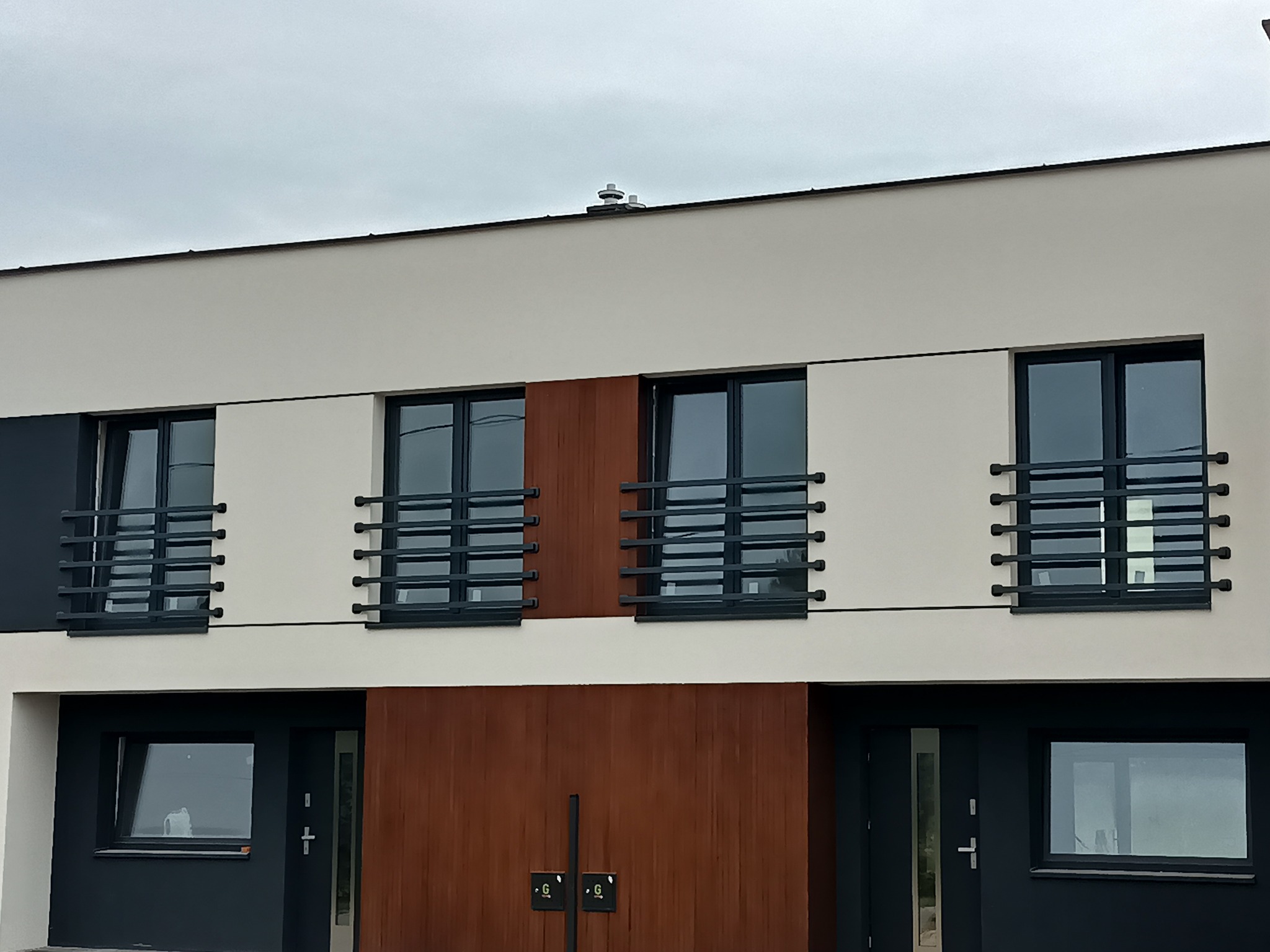 Nowoczesna fasada budynku z oknami zabezpieczonymi czarnymi, poziomymi kratami. Minimalistyczny design, neutralna kolorystyka ścian i drewniane akcenty.