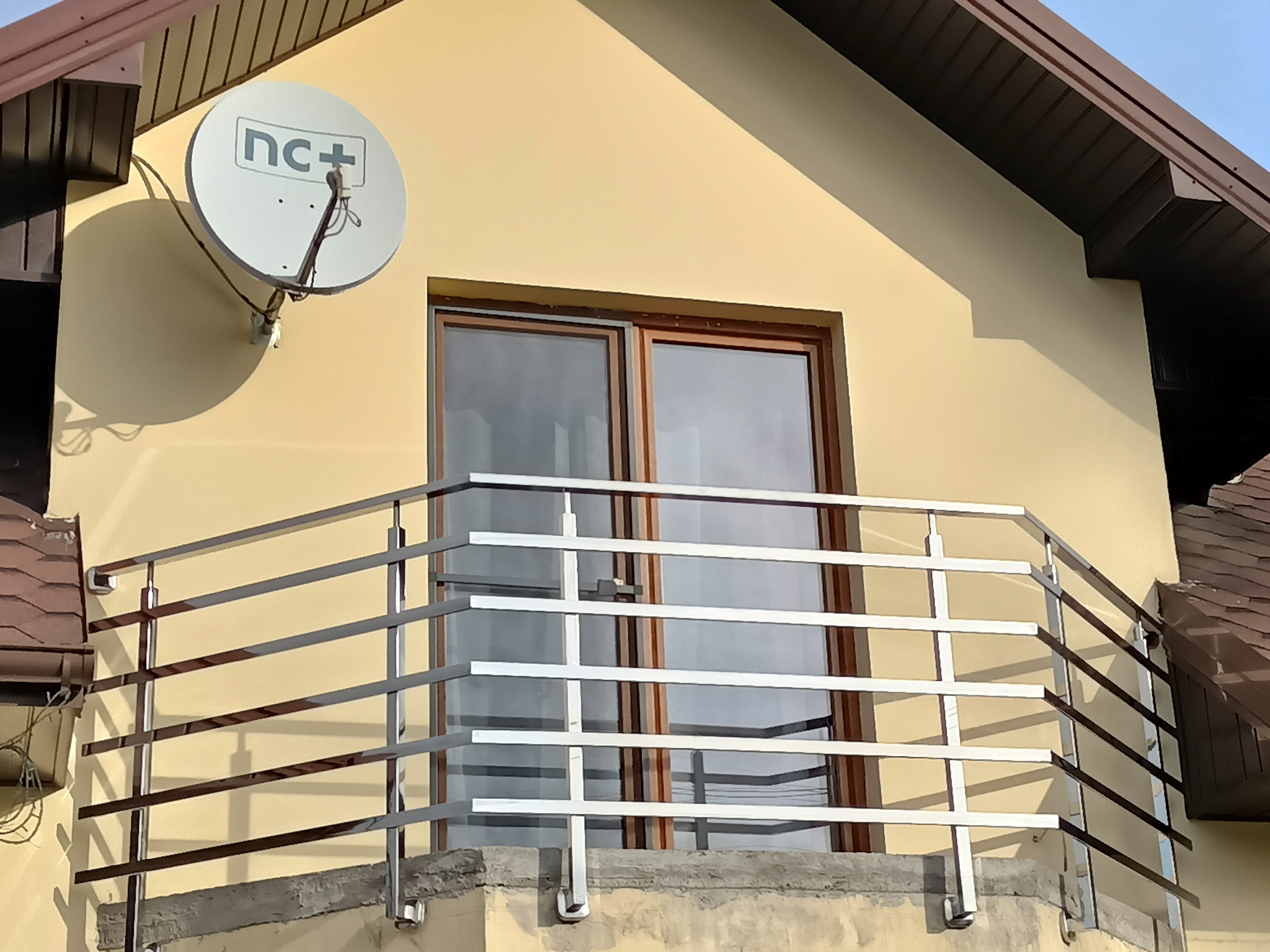 Balkon z nowoczesną, metalową balustradą. Na elewacji domu widoczna antena satelitarna. Ujęcie z dołu, podkreślające geometrię konstrukcji.