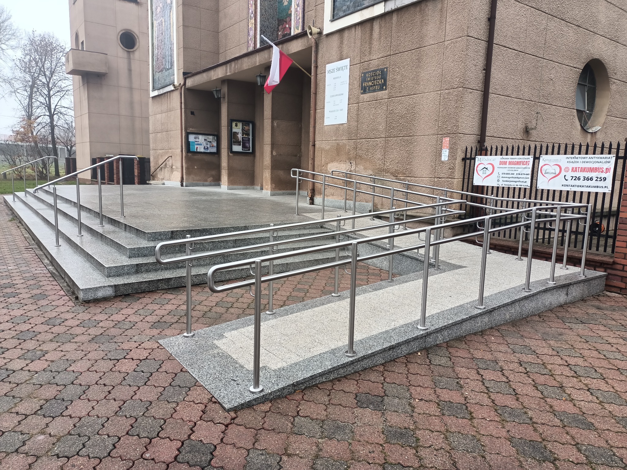 Zewnętrzne schody i rampa dla niepełnosprawnych z nierdzewnymi poręczami prowadzące do budynku kościoła. Widok na kostkę brukową i fragment elewacji.