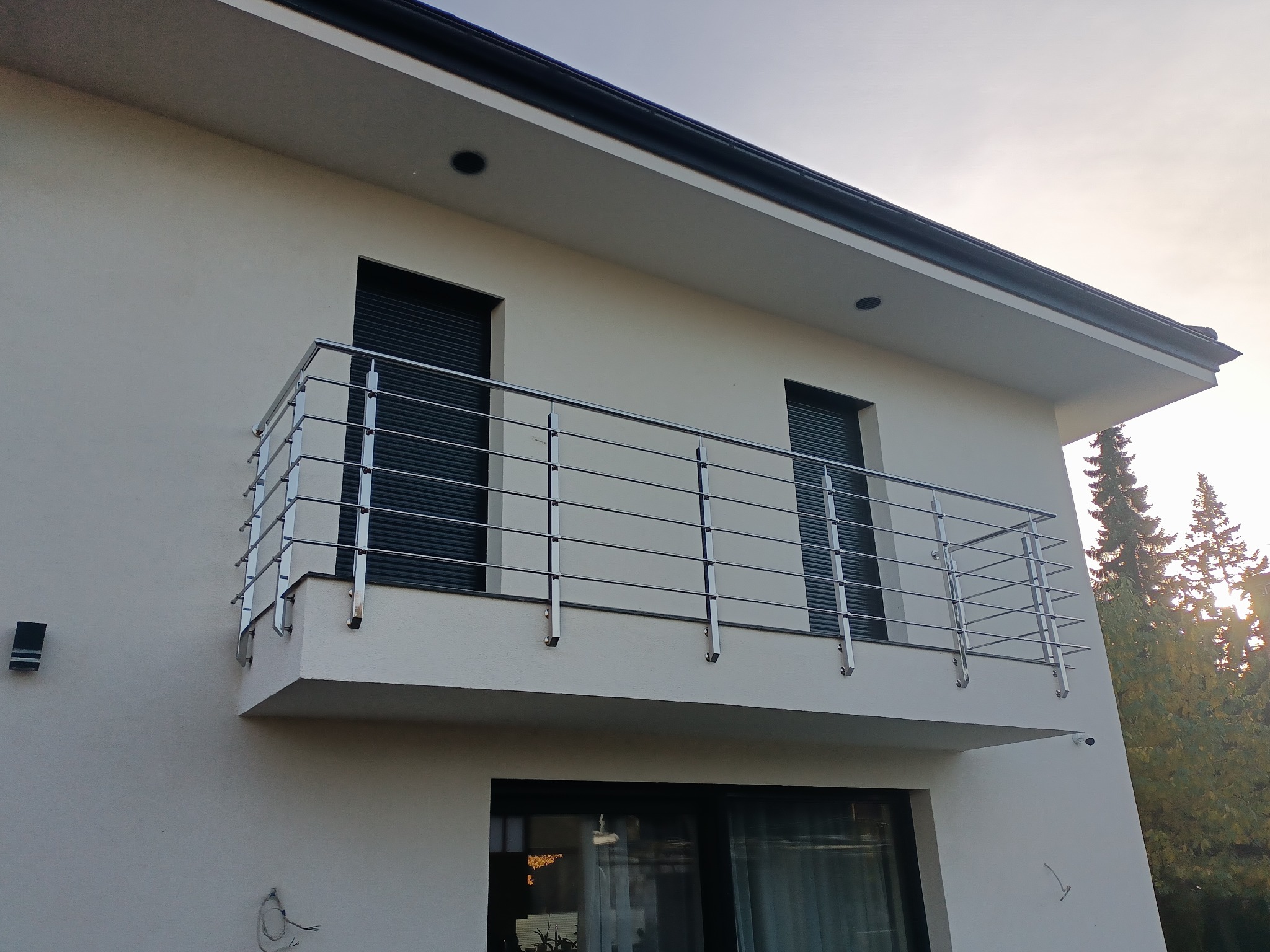 Balkon z nowoczesną, metalową balustradą na tle białej elewacji domu z roletami w oknach. Widok z dołu, podkreślający minimalistyczny design i symetrię konstrukcji.