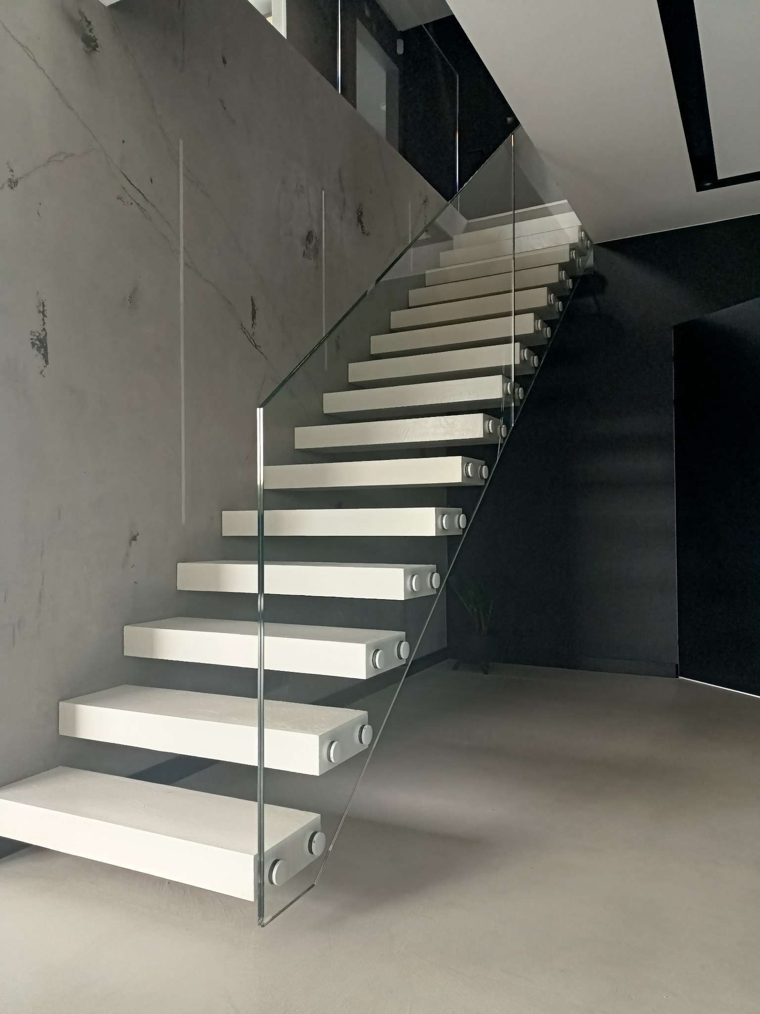 Nowoczesne, minimalistyczne schody z białymi stopniami i szklaną balustradą na tle szarej ściany z efektem betonu, widok z dołu, oświetlenie naturalne.