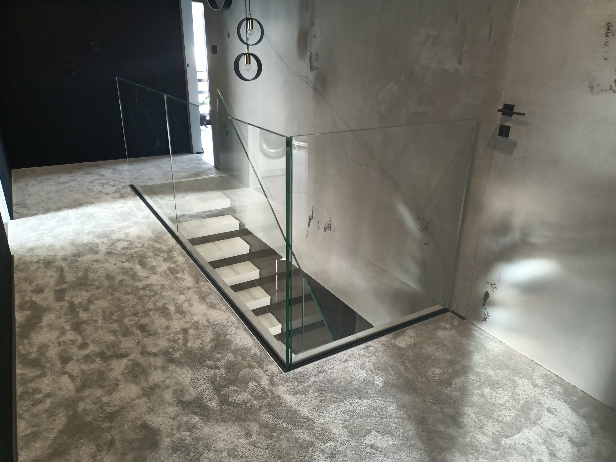 Nowoczesne schody z przeszkloną balustradą w minimalistycznym wnętrzu. Szklane panele z czarnymi detalami podkreślają surowy charakter wykończenia ścian i podłogi.