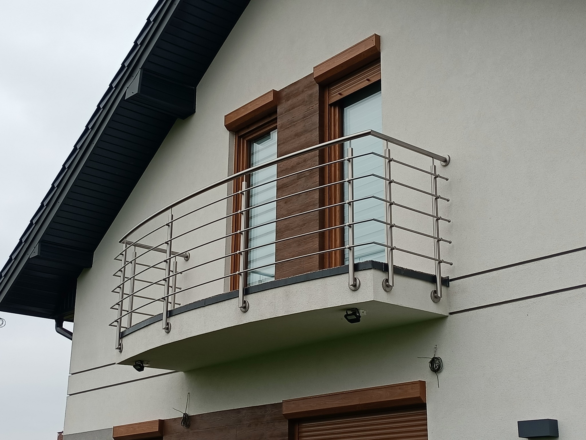 Nowoczesny balkon z balustradą ze stali nierdzewnej na tle jasnej elewacji domu z drewnianymi akcentami okiennymi i czarnym dachem. Ujęcie z dołu.