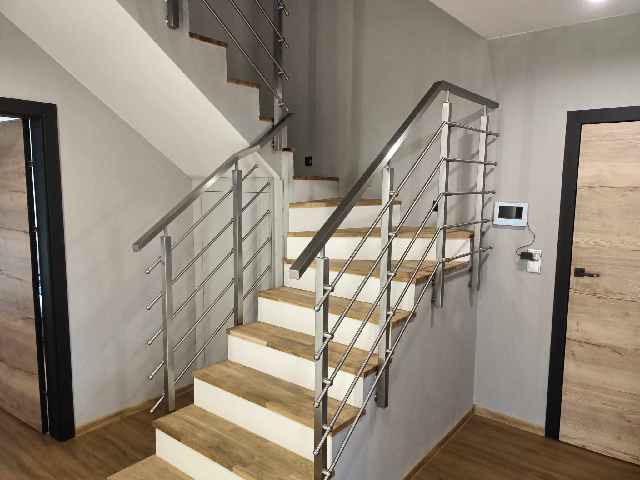 Nowoczesne schody z drewnianymi stopniami i metalową balustradą w minimalistycznym wnętrzu. Widoczny fragment drzwi i panel sterowania na ścianie.