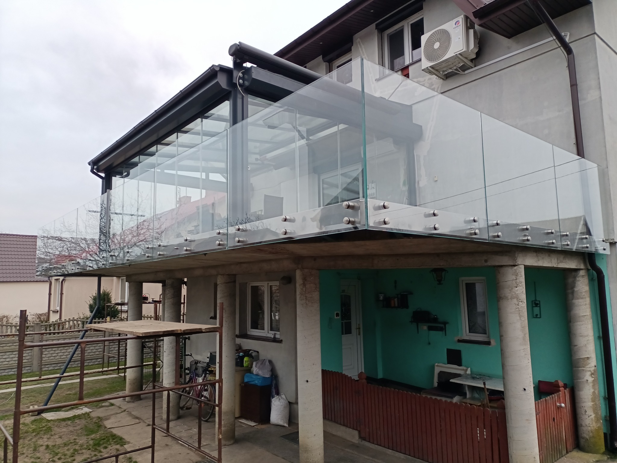 Szklana balustrada balkonu z widocznymi mocowaniami punktowymi, nowoczesna konstrukcja na tle domu z klimatyzacją. Widok z dołu, w tle drzewa bez liści.