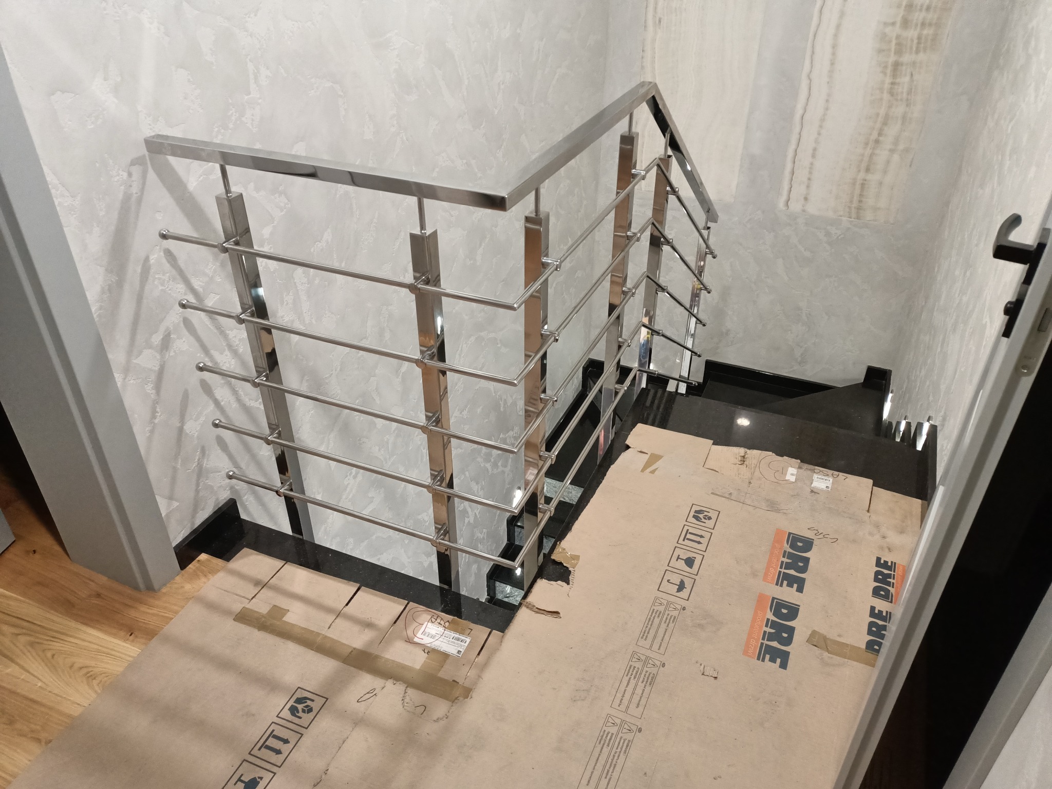 Nowoczesne, polerowane poręcze ze stali nierdzewnej zamontowane na czarnych schodach z marmuru, zabezpieczone kartonem. Ściany wykończone dekoracyjnym tynkiem.