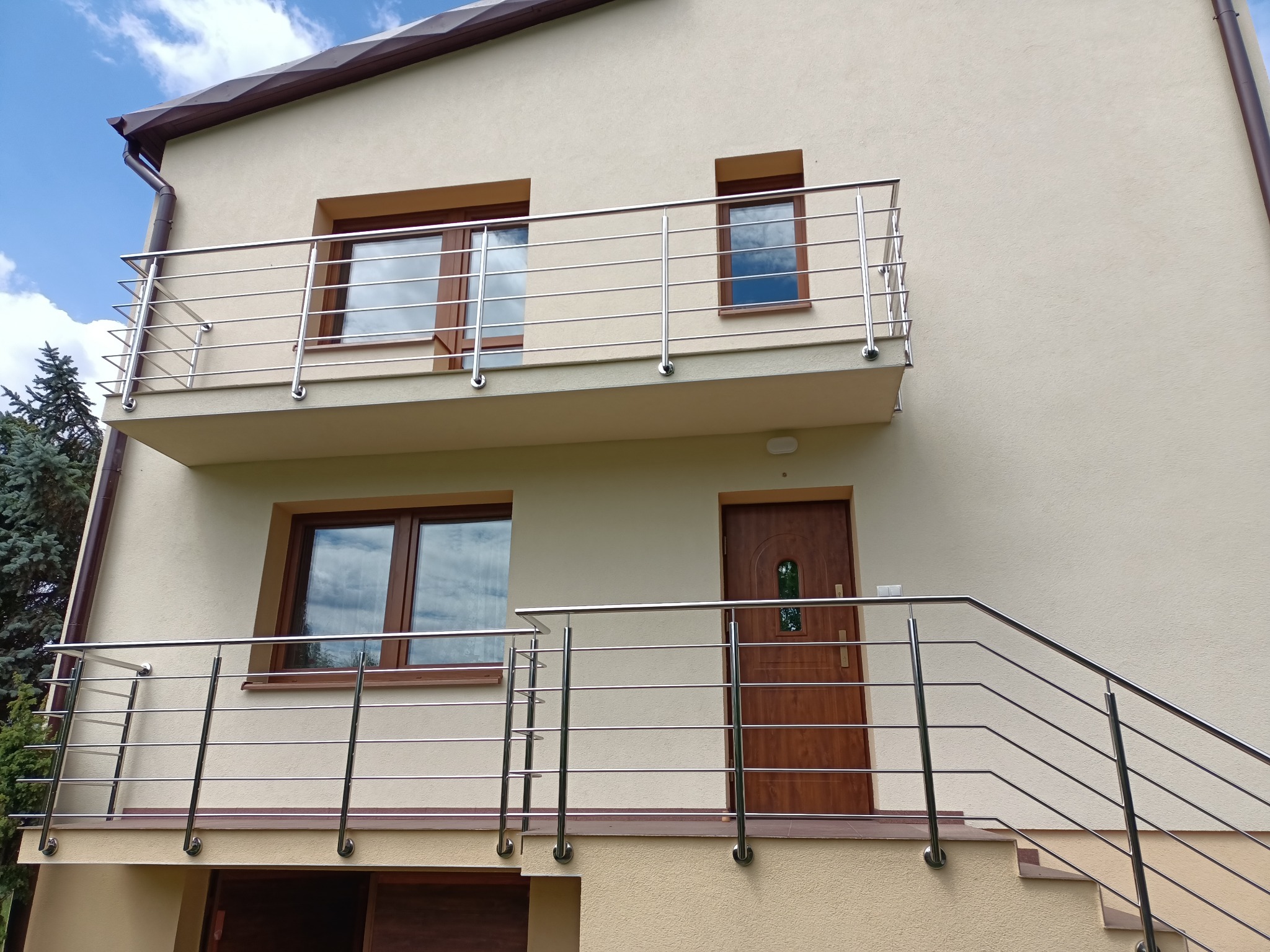 Dwukondygnacyjny dom z beżową elewacją. Balkon i schody wejściowe z metalową, poziomą balustradą, brązowe okna i drzwi. Niebieskie niebo w tle.