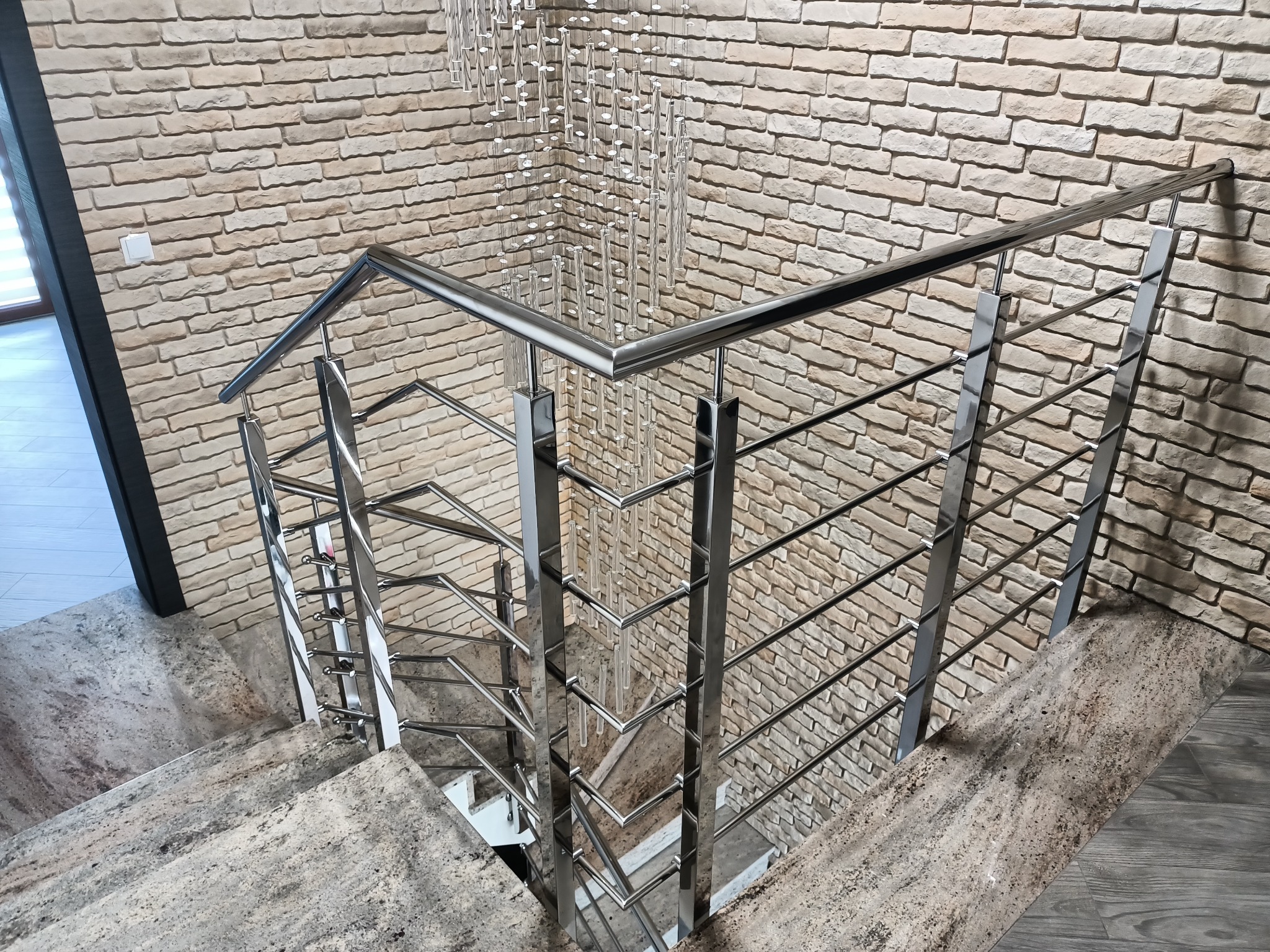 Polerowana, metalowa balustrada schodowa z wypełnieniem z prętów, na tle ściany z cegły i kryształowego żyrandola, widziana z góry.