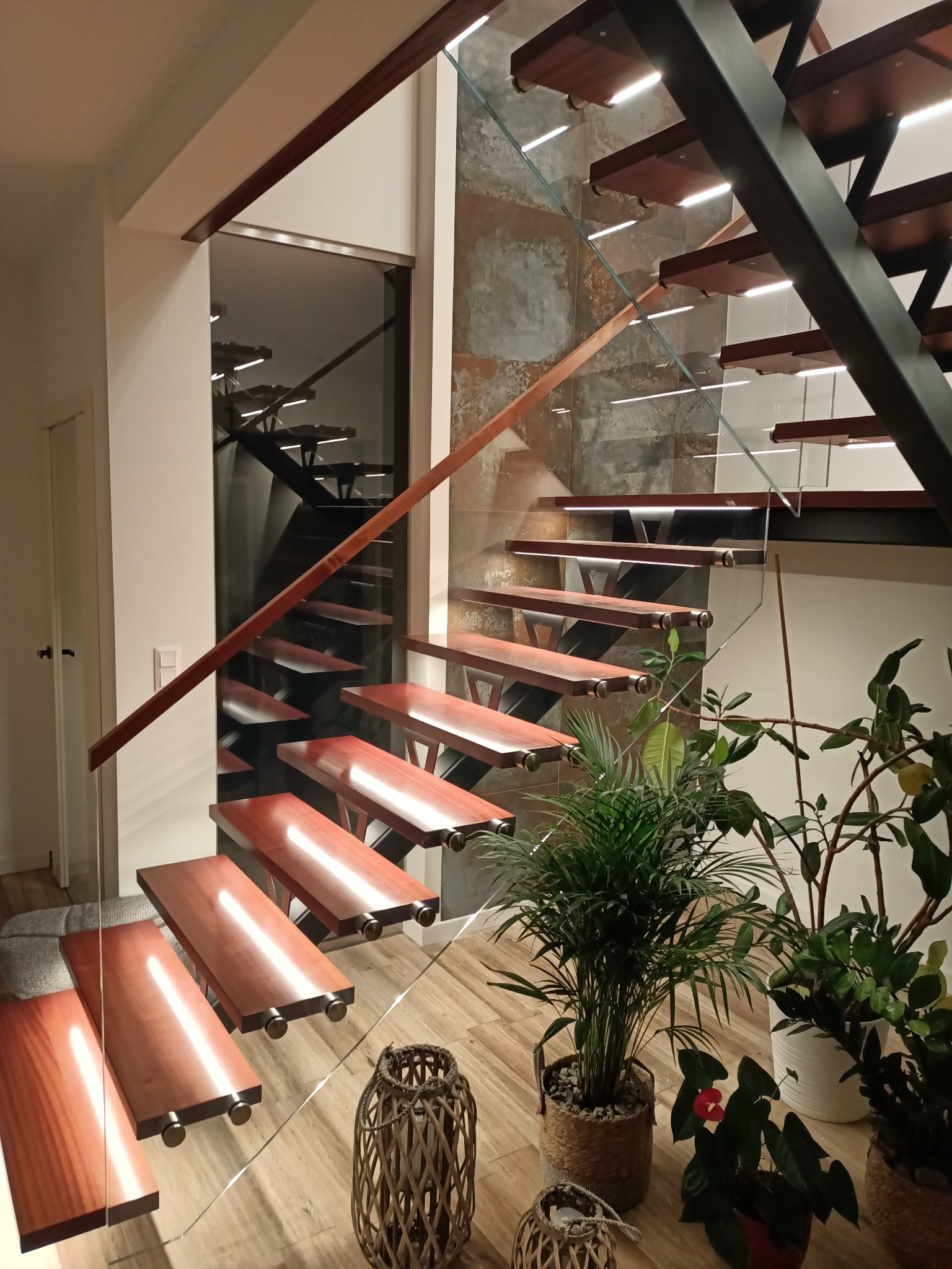 Nowoczesne schody drewniane z podświetleniem i szklaną balustradą, widoczne rośliny doniczkowe, tworzące przytulny kącik w domu. Ujęcie z dołu, podkreślające lekkość konstrukcji.
