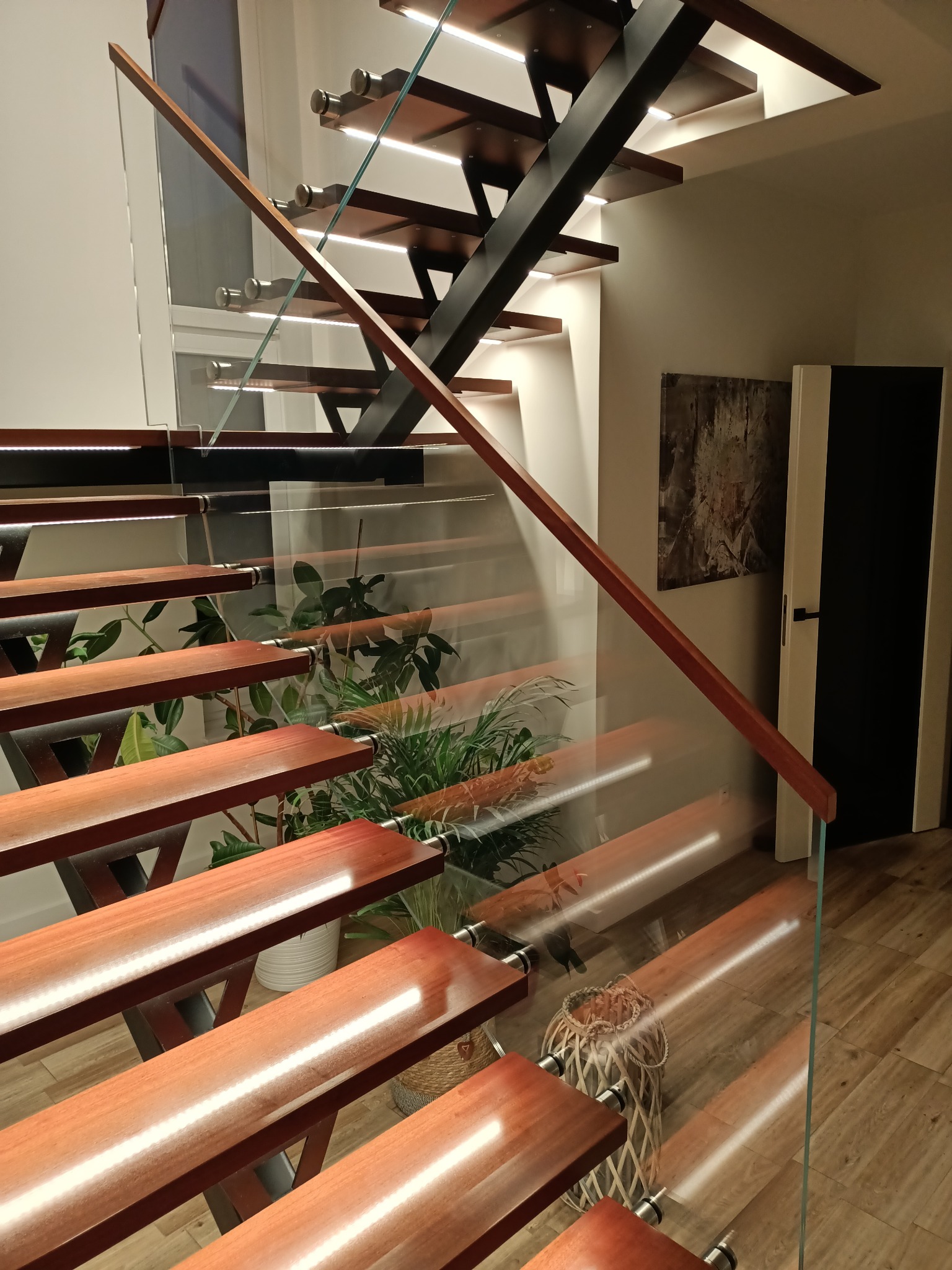 Nowoczesne schody metalowe z drewnianymi stopniami i szklaną balustradą, podświetlane LED. Widoczne rośliny doniczkowe i grafika na ścianie.